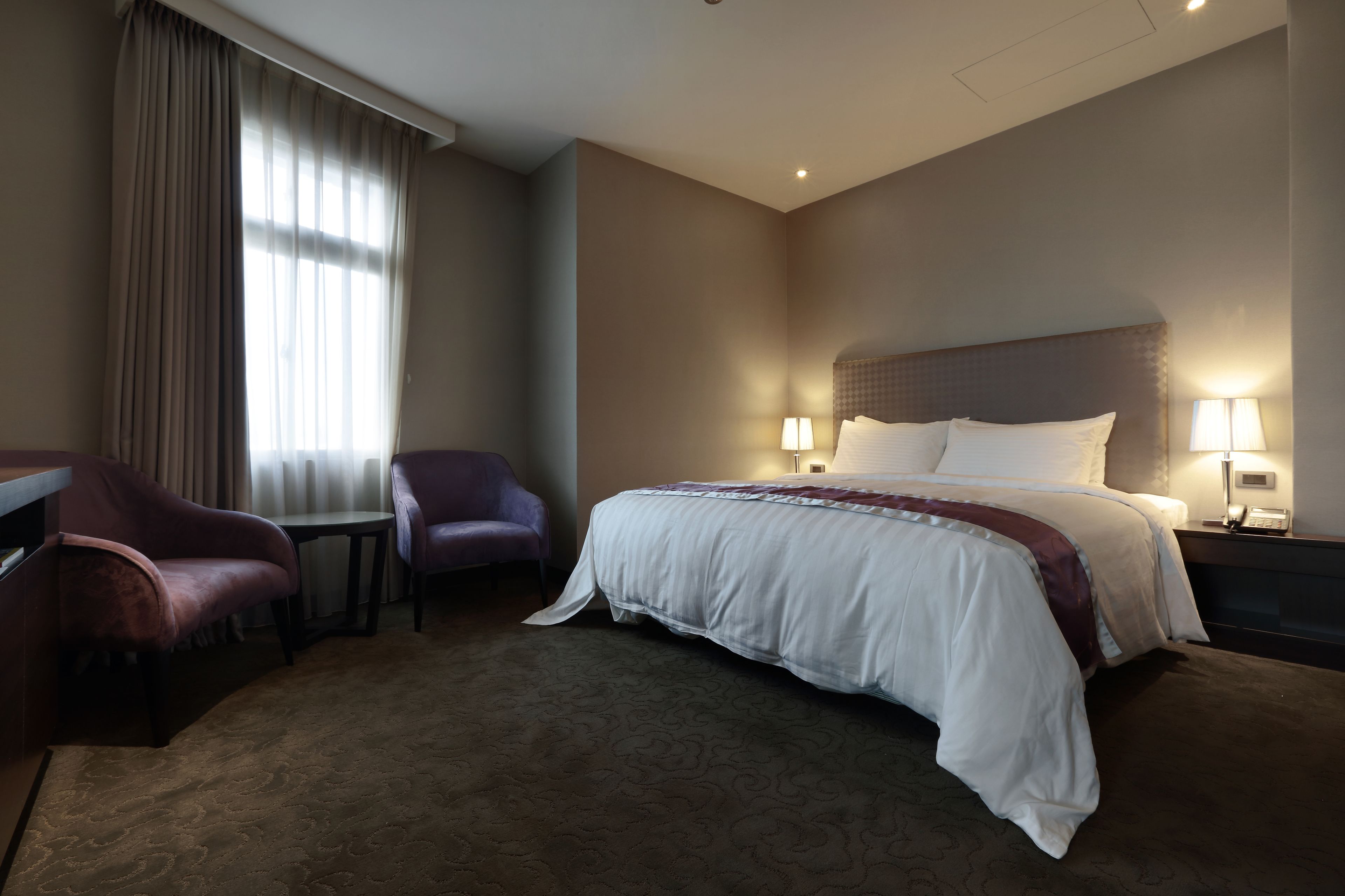 A hotel room with a large bed, two bedside lamps, a desk, a chair, and a window with curtains.