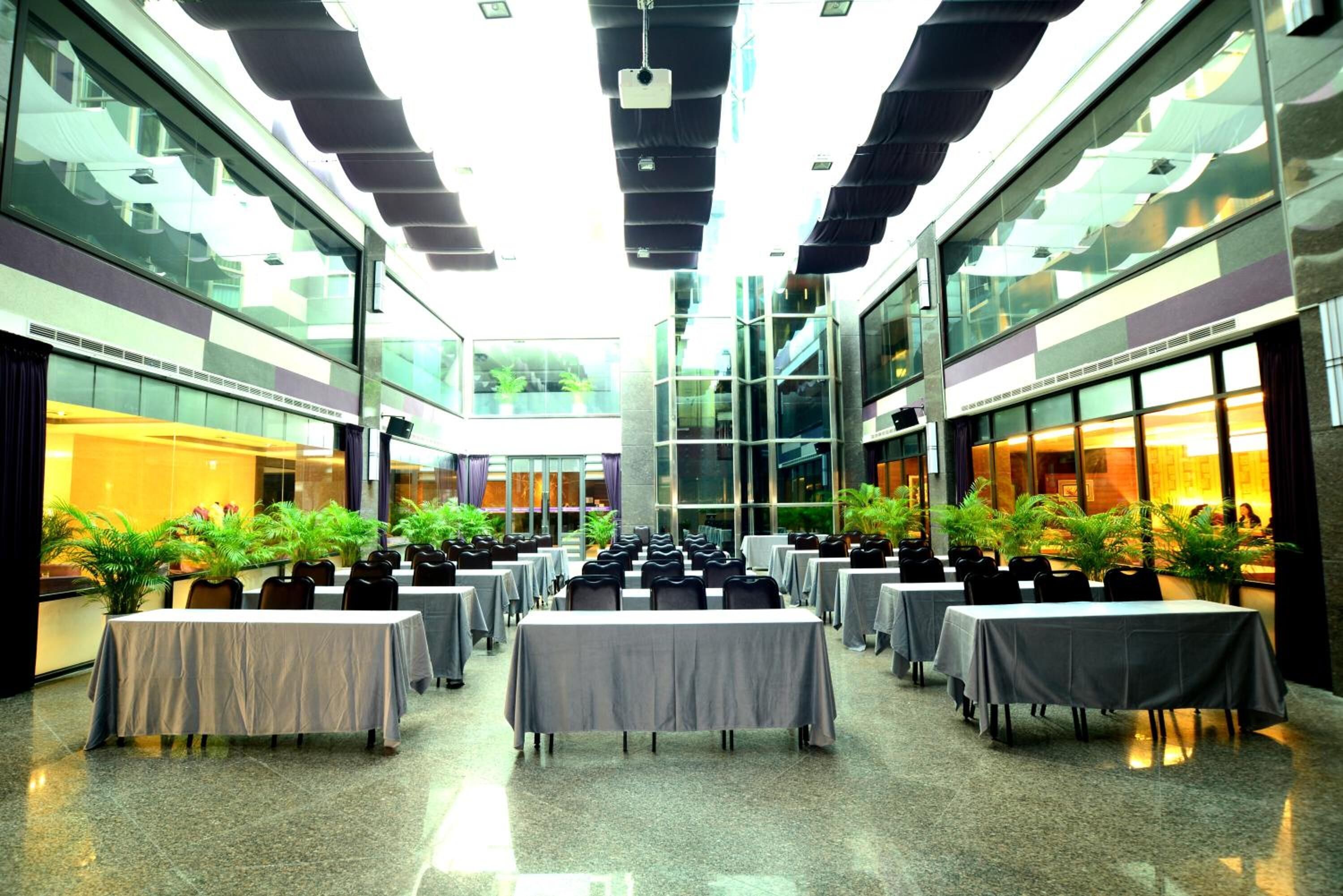 A spacious indoor area with multiple tables and chairs arranged in rows, large windows on both sides, and potted plants.