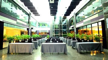 A spacious indoor area with multiple tables and chairs arranged in rows, large windows on both sides, and potted plants.