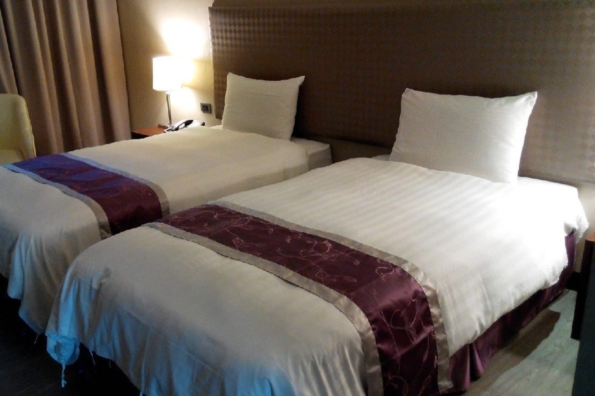 A hotel room with two beds, a chair, a lamp, and a curtain.