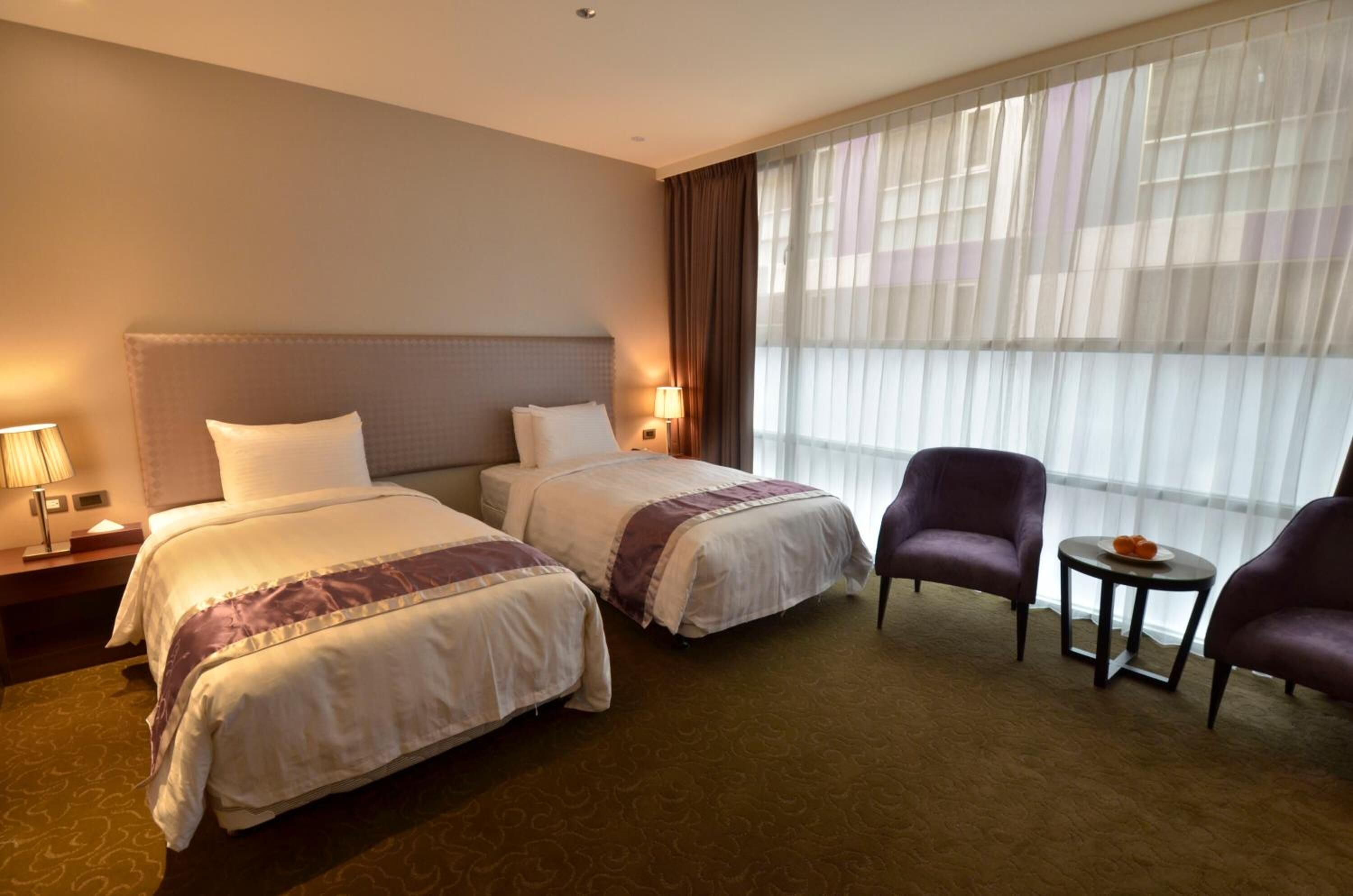 A hotel room with two beds, a sitting area with a chair and a small table, and a large window with sheer curtains.