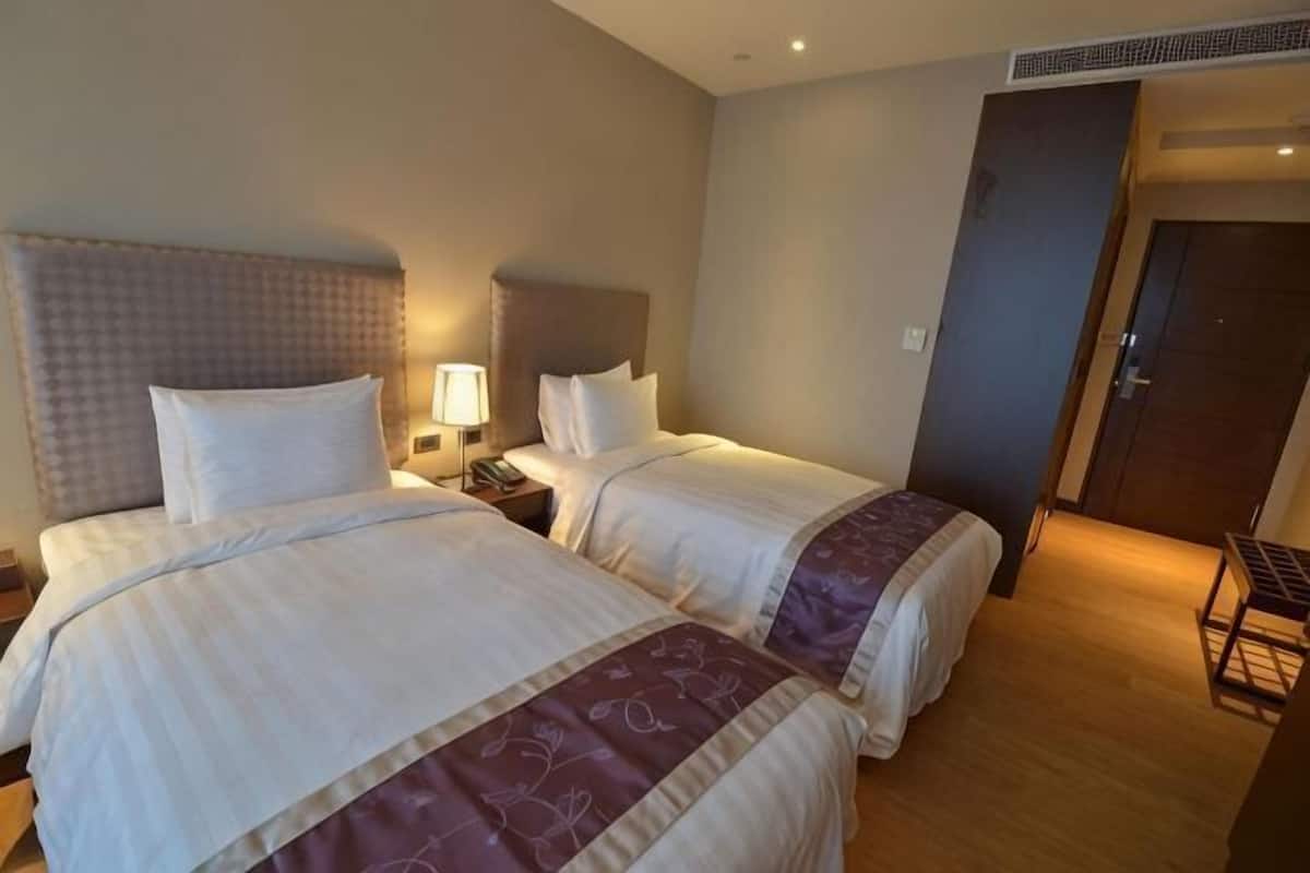 A hotel room with two beds, a nightstand with a lamp, a wooden floor, and a door leading to another room.