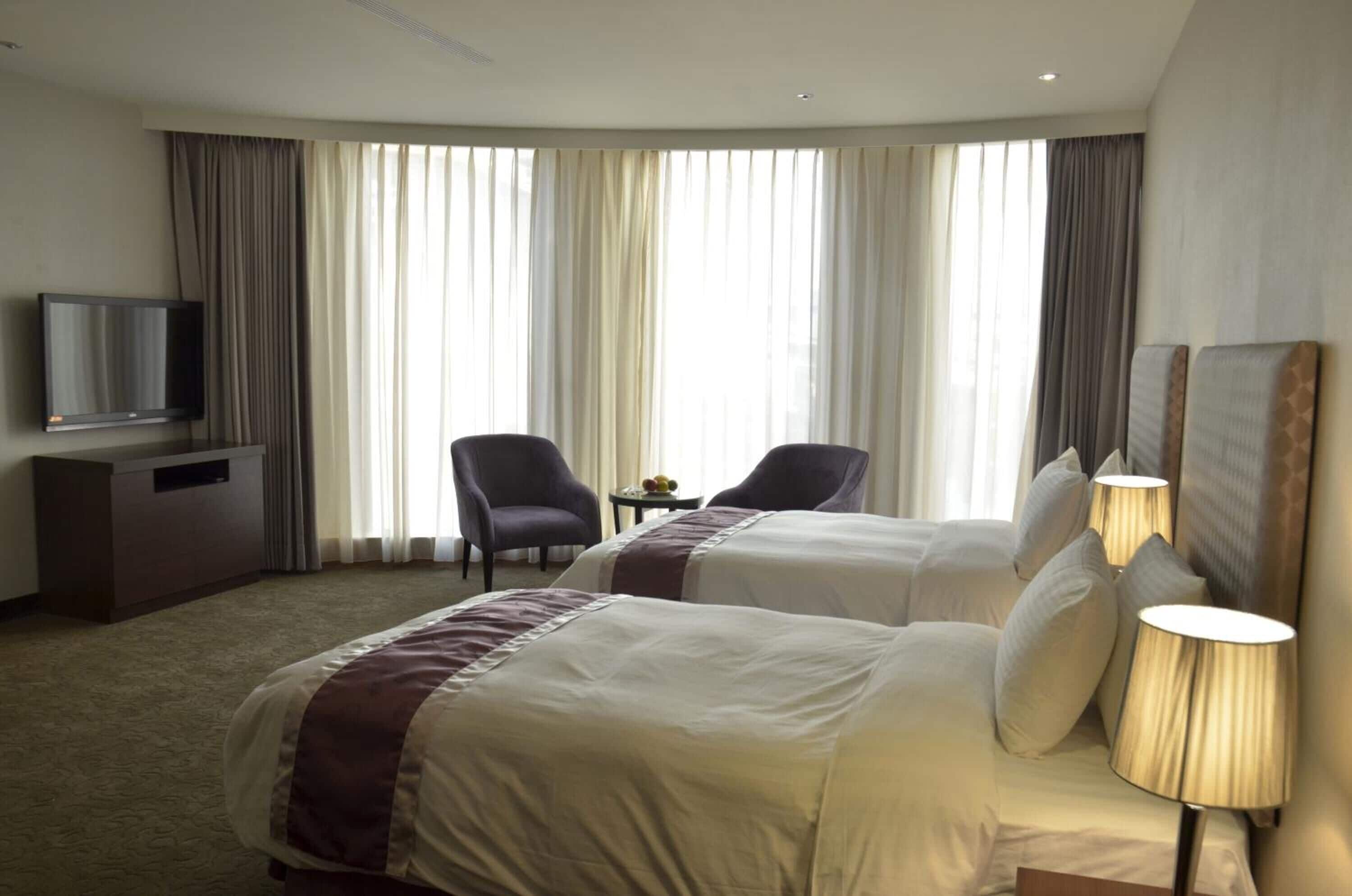 A hotel room with two beds, a TV, a desk, and a view of the city through large windows.