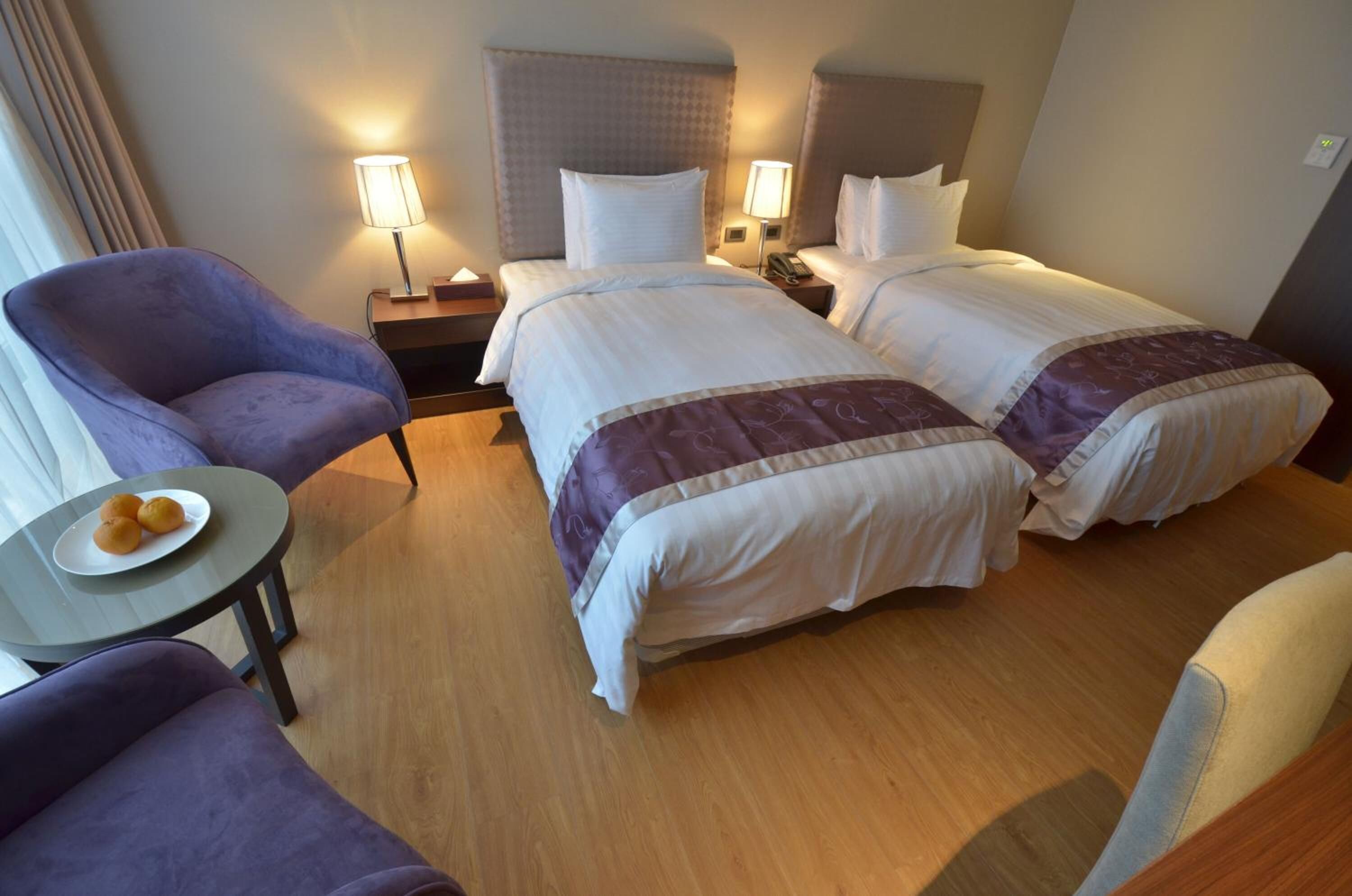 A hotel room with two beds, a small table with a plate of fruit, and a purple armchair.