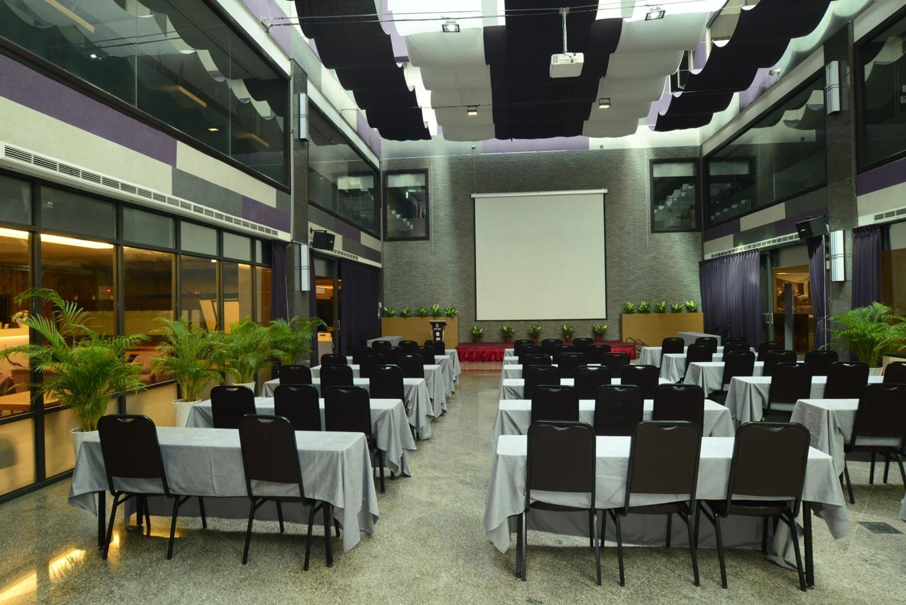 A conference room with multiple tables and chairs, a large screen, and potted plants.