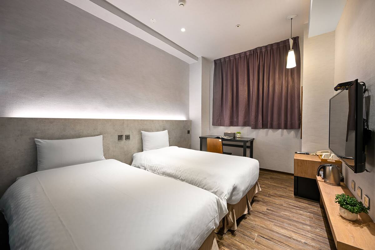 A modern hotel room with two beds, a desk, a TV, and a window with curtains.