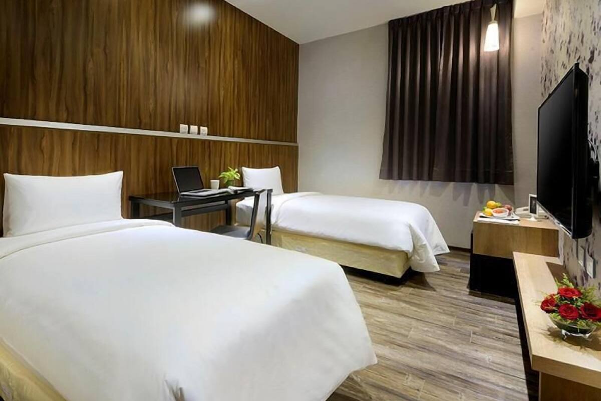 A hotel room with two beds, a desk with a laptop, a TV, and a window with curtains.