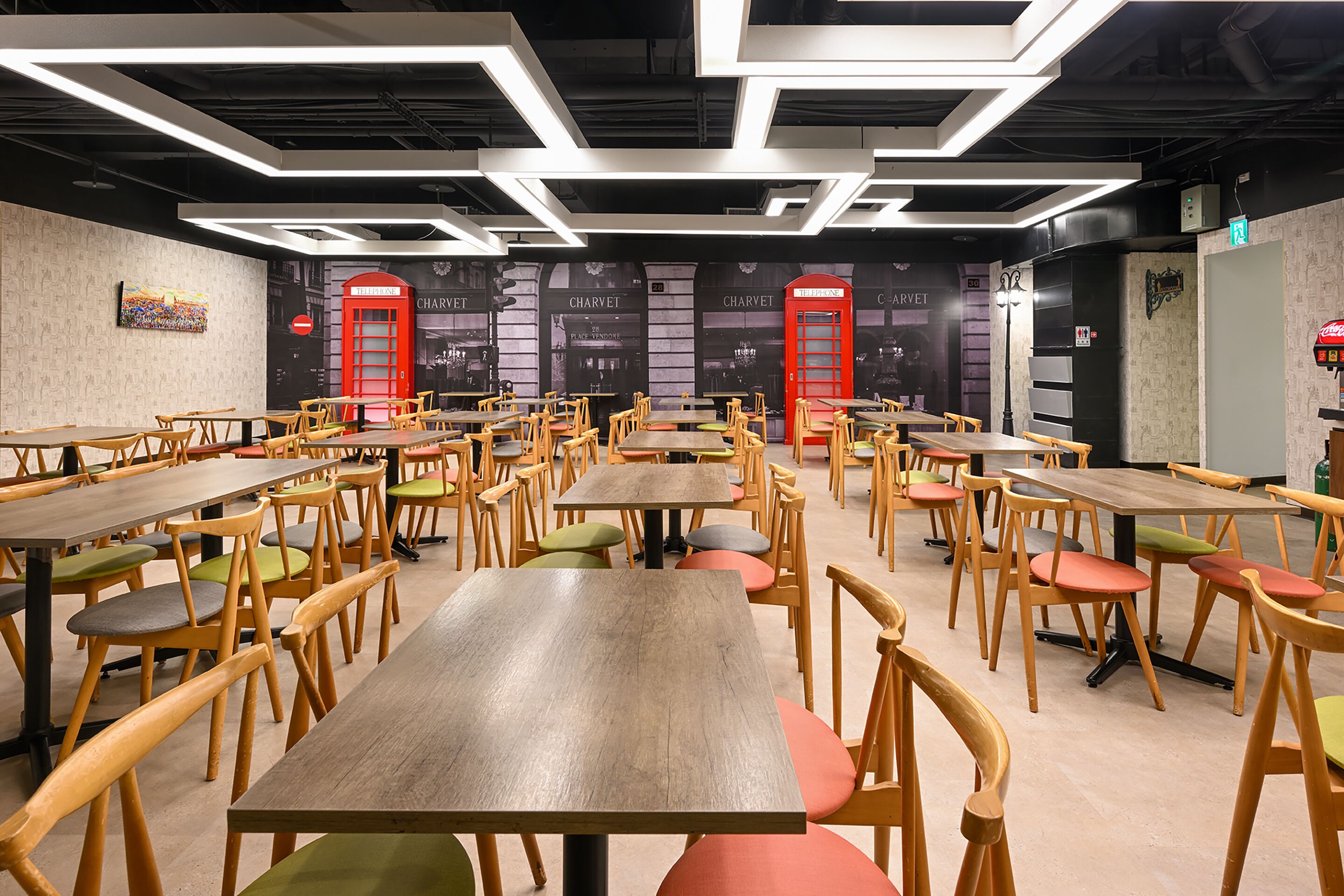 A spacious dining area with multiple wooden tables and chairs, featuring a modern ceiling design and red doorways.