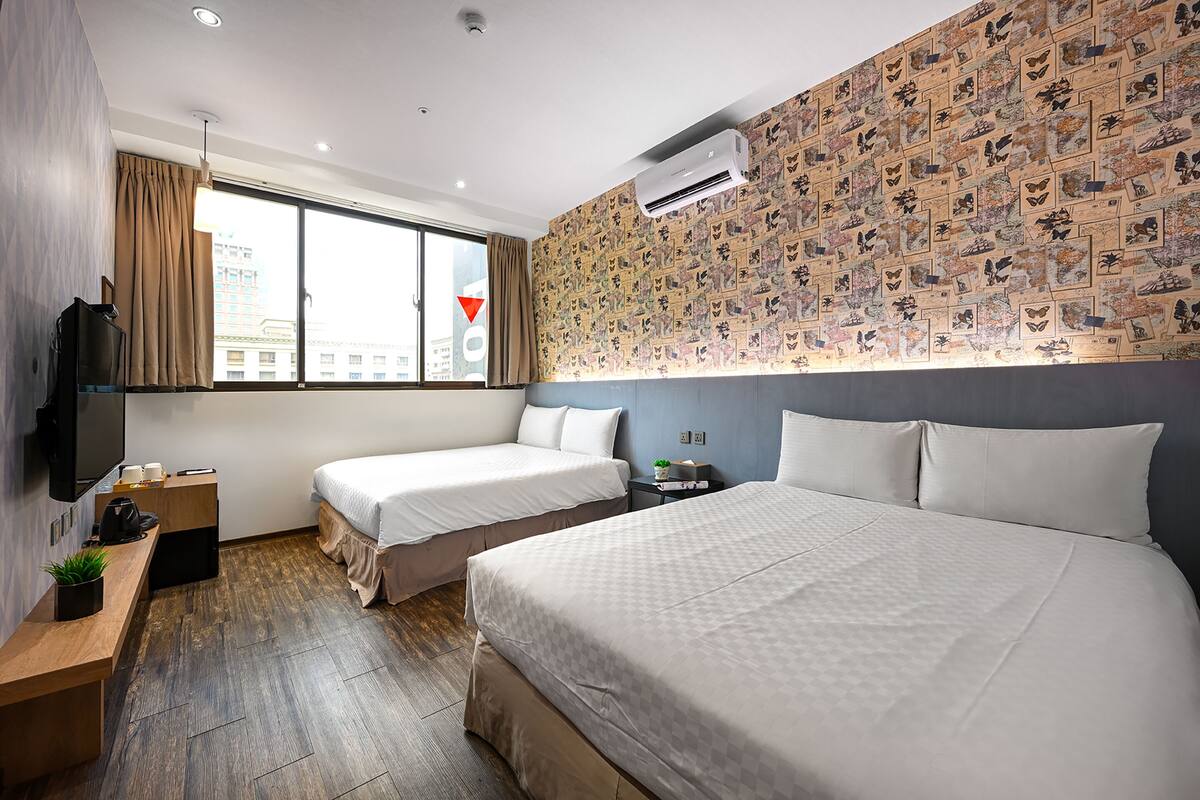A hotel room with two beds, a TV, a desk, and a large window with curtains.