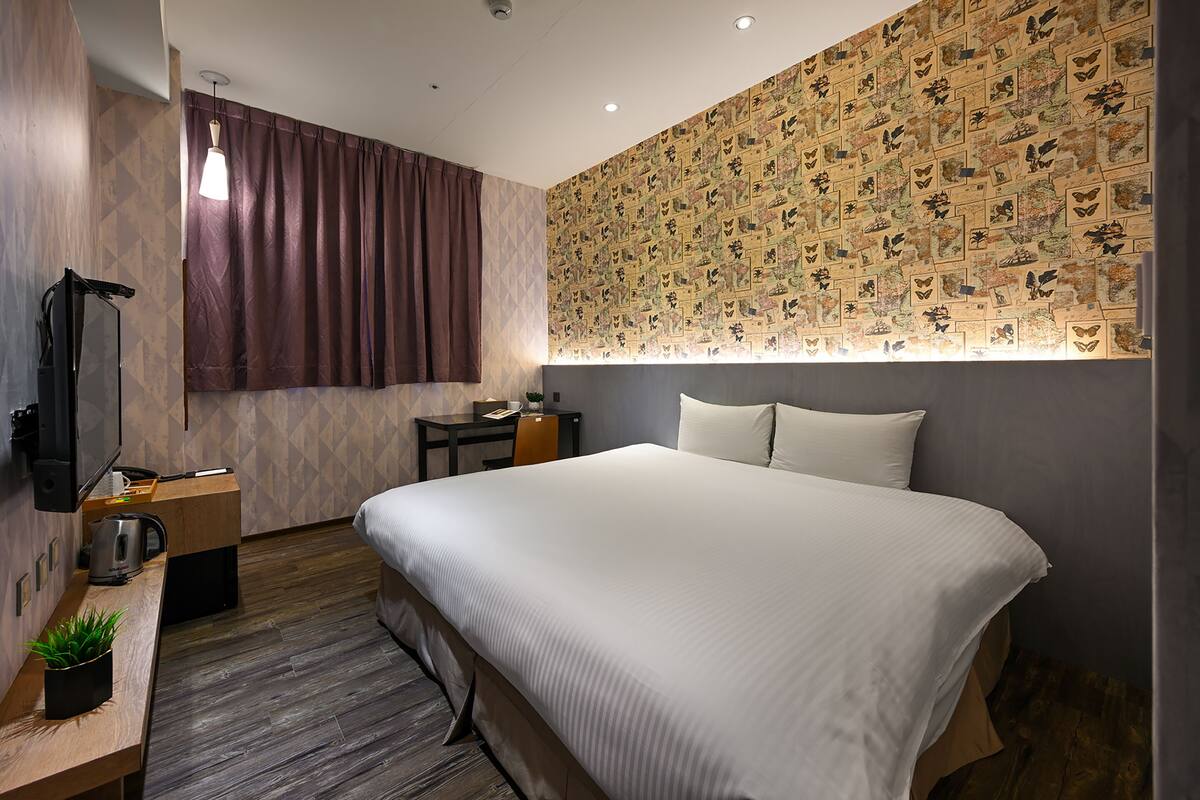 A modern hotel room with a large bed, a desk with a kettle, a TV, and a wall with a butterfly pattern.