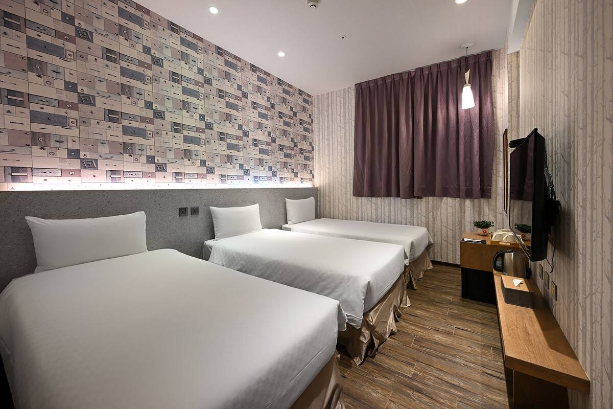 A hotel room with three beds, a wooden desk, a television, and a patterned wall.
