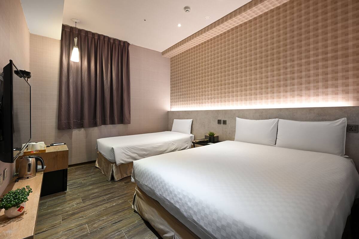A hotel room with two beds, a desk with a kettle, a small plant, and a wall-mounted TV.