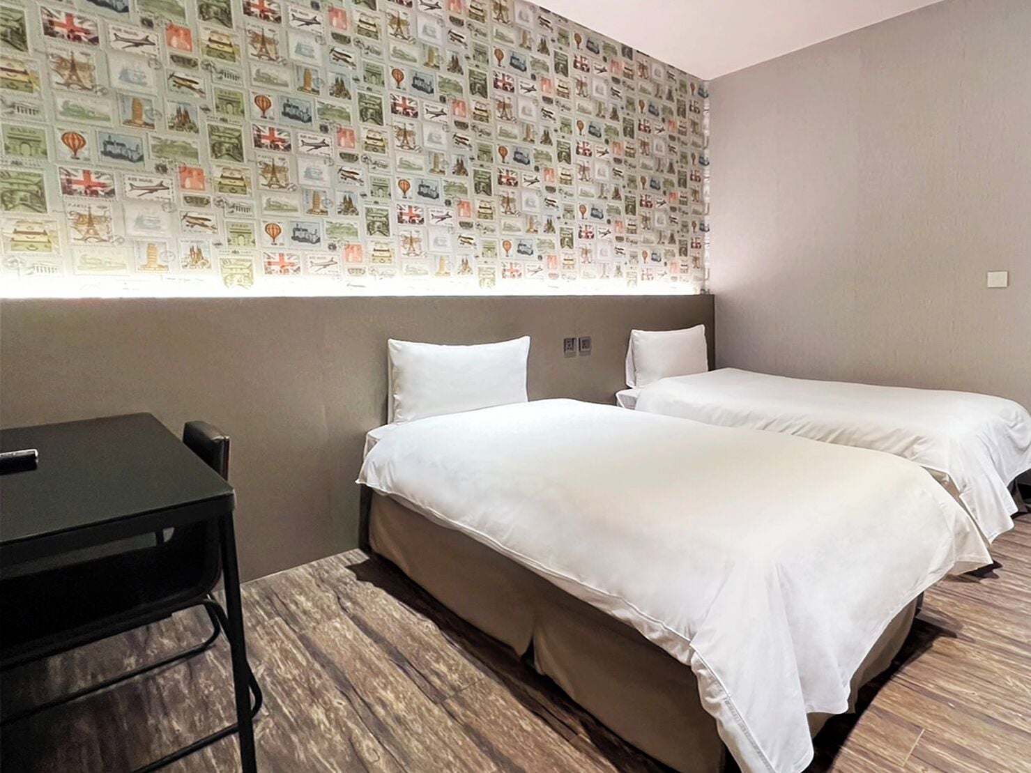 Une chambre d’hôtel avec deux lits, un bureau et un mur décoré de cartes postales sur le thème du voyage.