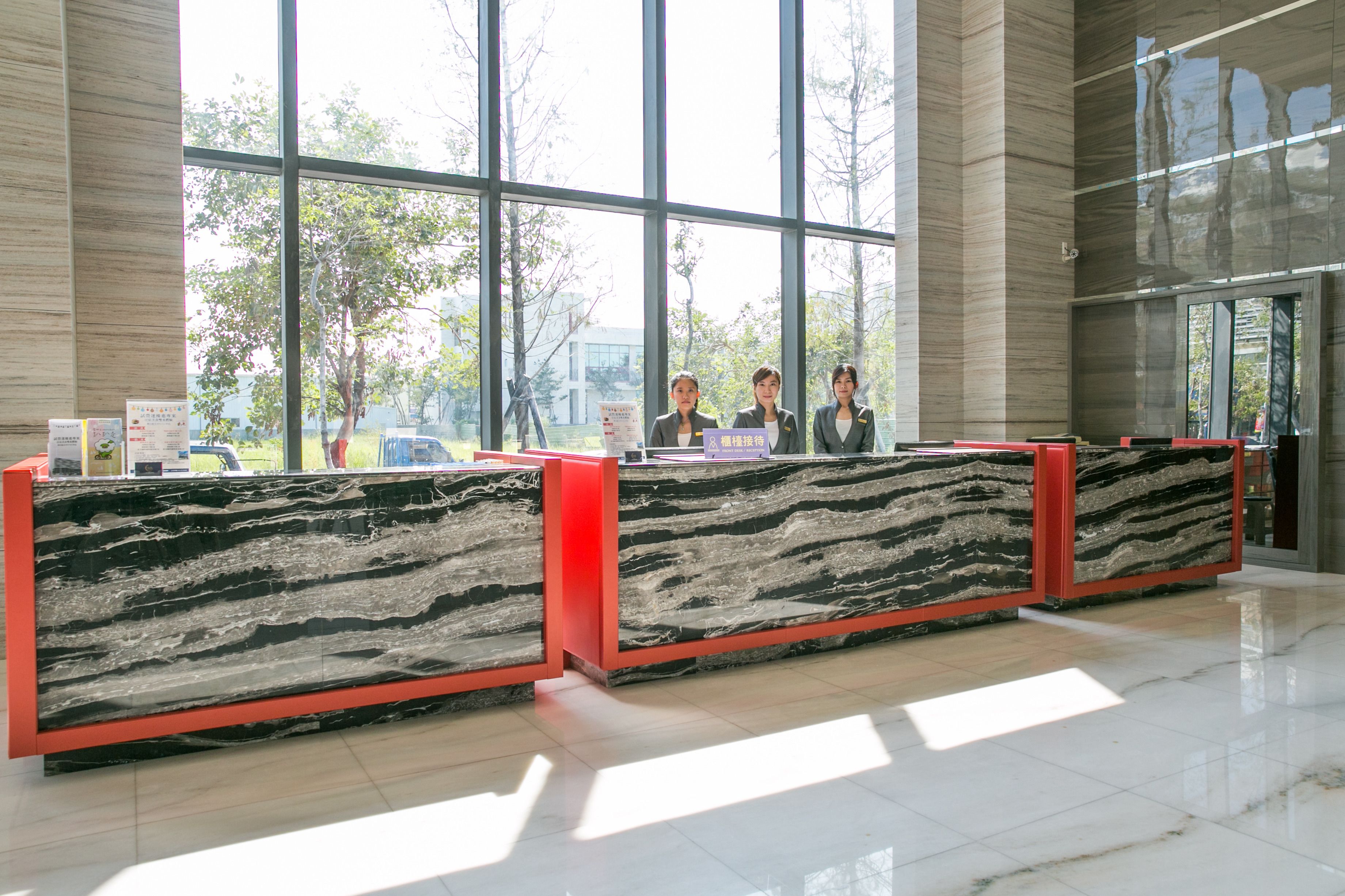 A reception desk with a marble top and red base, situated in a spacious lobby with large windows and a tiled floor.