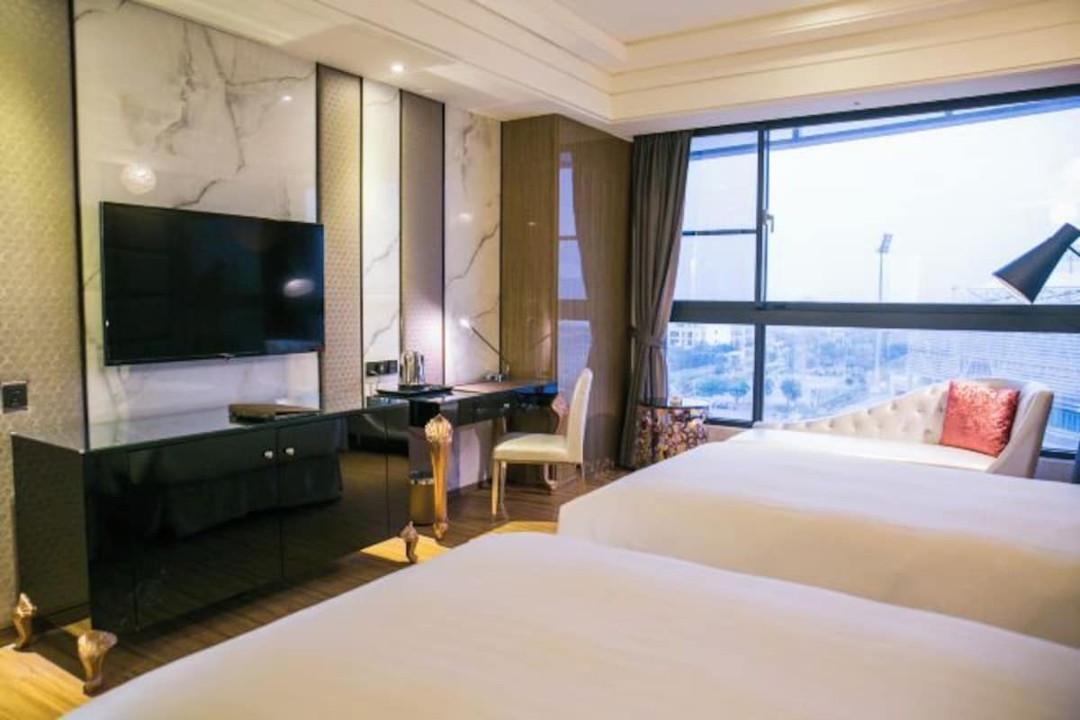 A hotel room with a large bed, a desk with a computer, a flat-screen TV, and a large window offering a city view.