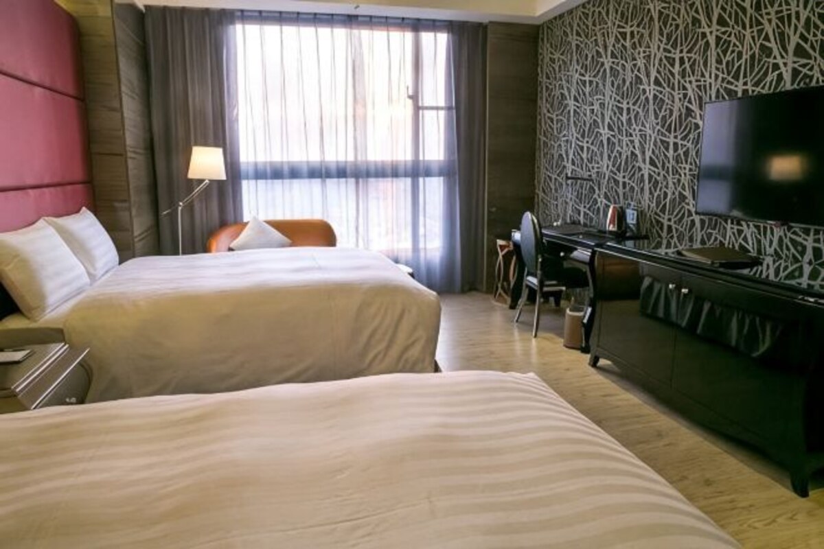 A hotel room with two beds, a desk, a chair, a TV, and a large window with curtains.