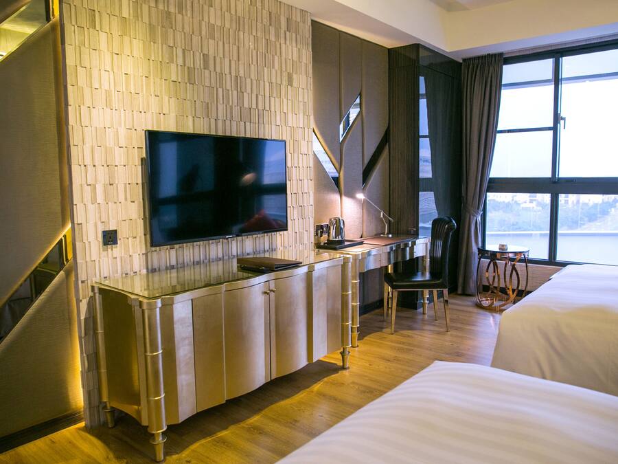 A hotel room with a large flat-screen TV mounted on a wall, a desk with a chair, a bed, and a view of the outdoors through a large window.