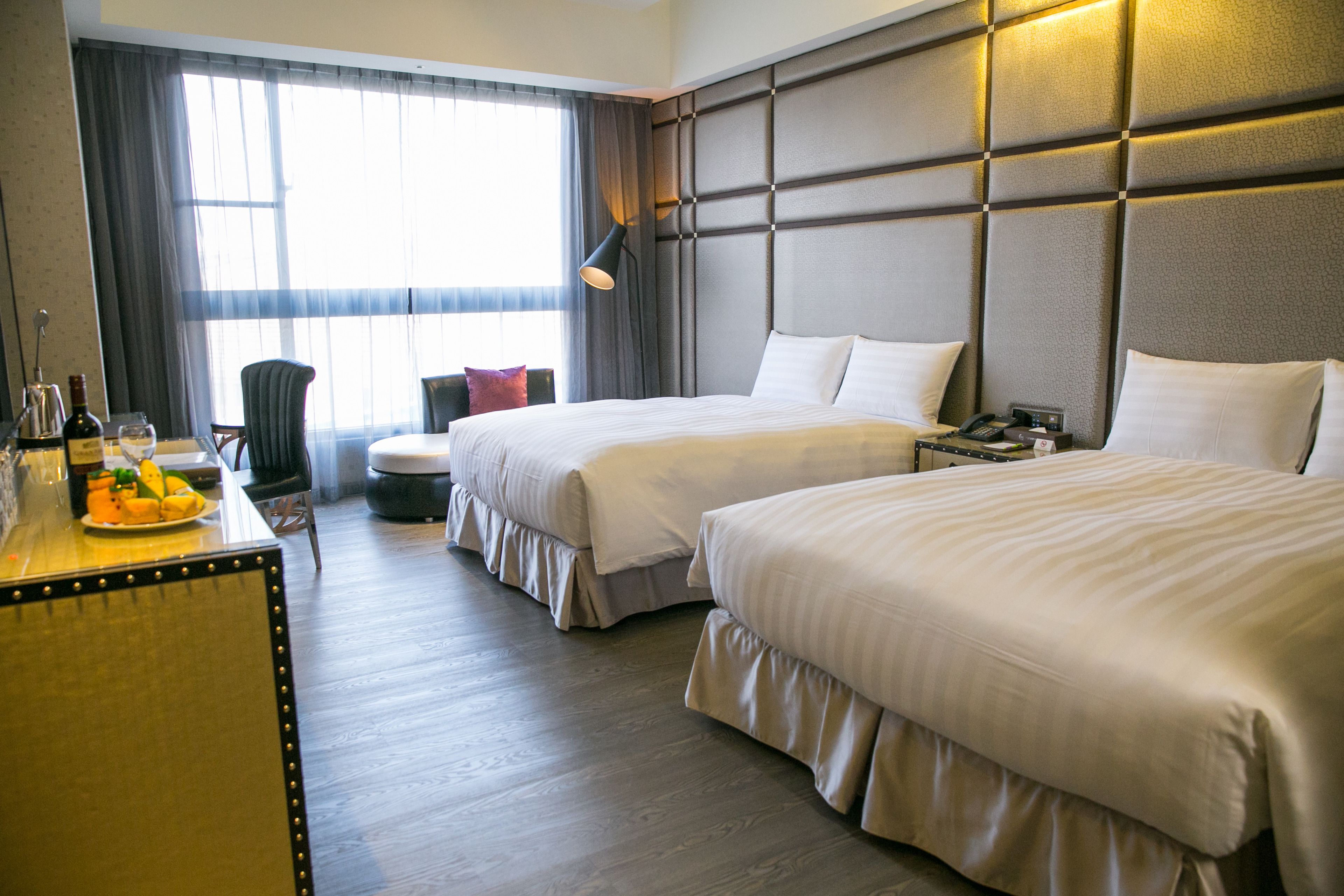 Habitación de hotel con dos camas, un escritorio con lámpara, una silla y un ventanal con cortinas.