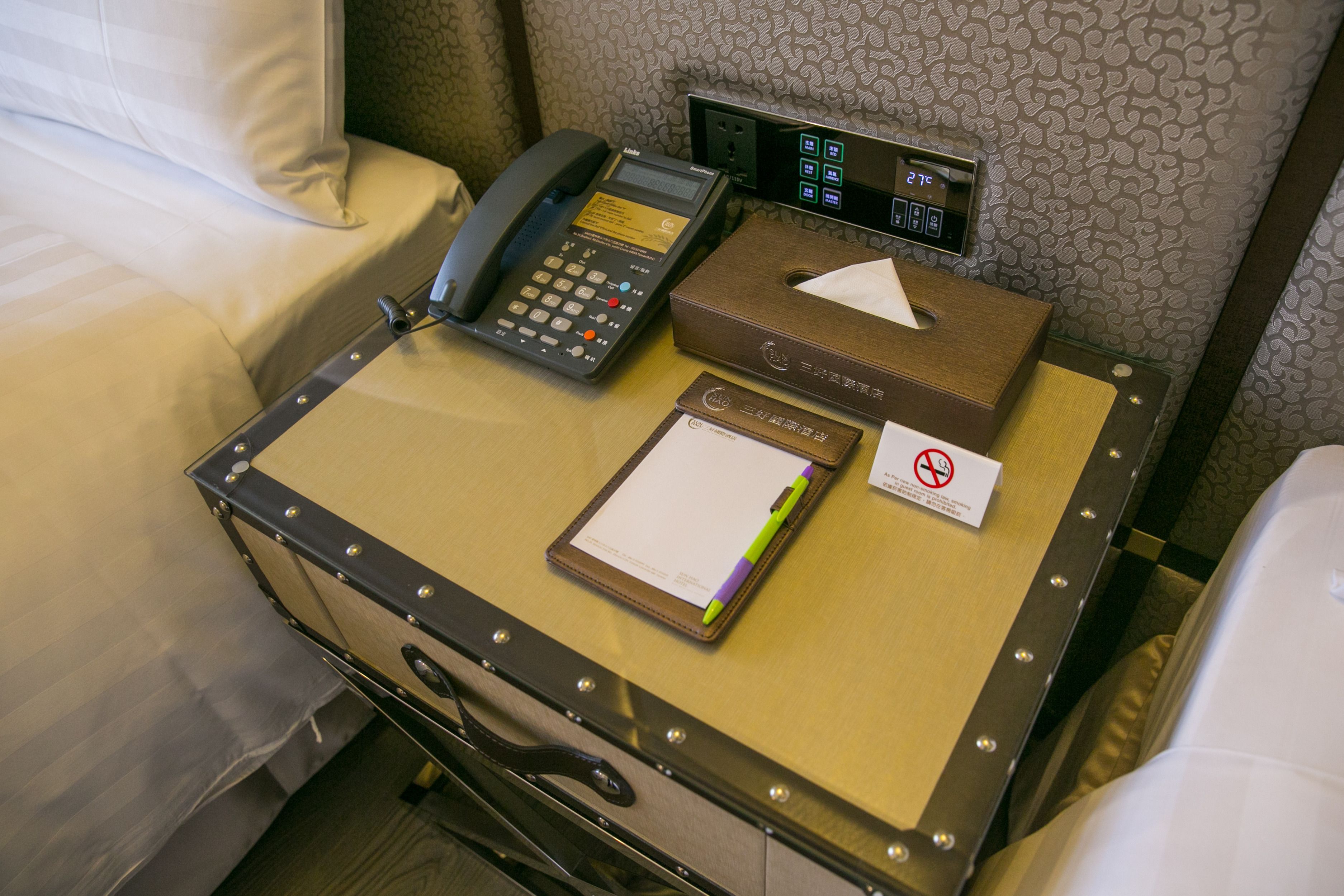 A hotel room with a bedside table containing a telephone, a tissue box, a notepad, and a pen.