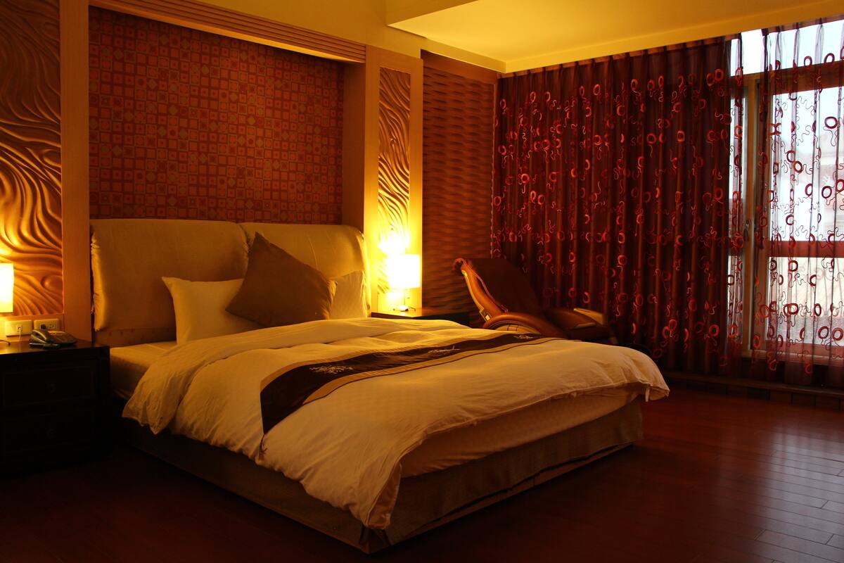 A large bed with a headboard, two bedside lamps, and a chair in a warmly lit room with patterned wallpaper and curtains.