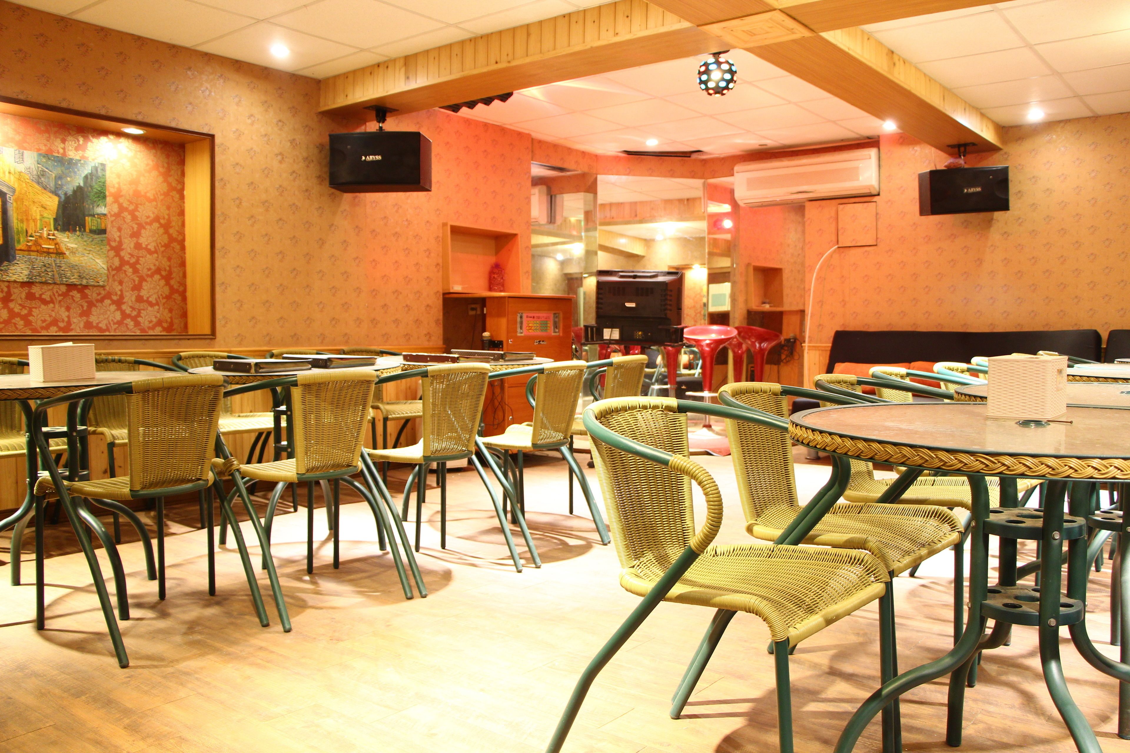 Un coin repas avec des chaises et des tables en osier, un grand tableau accroché au mur et deux téléviseurs fixés au mur.