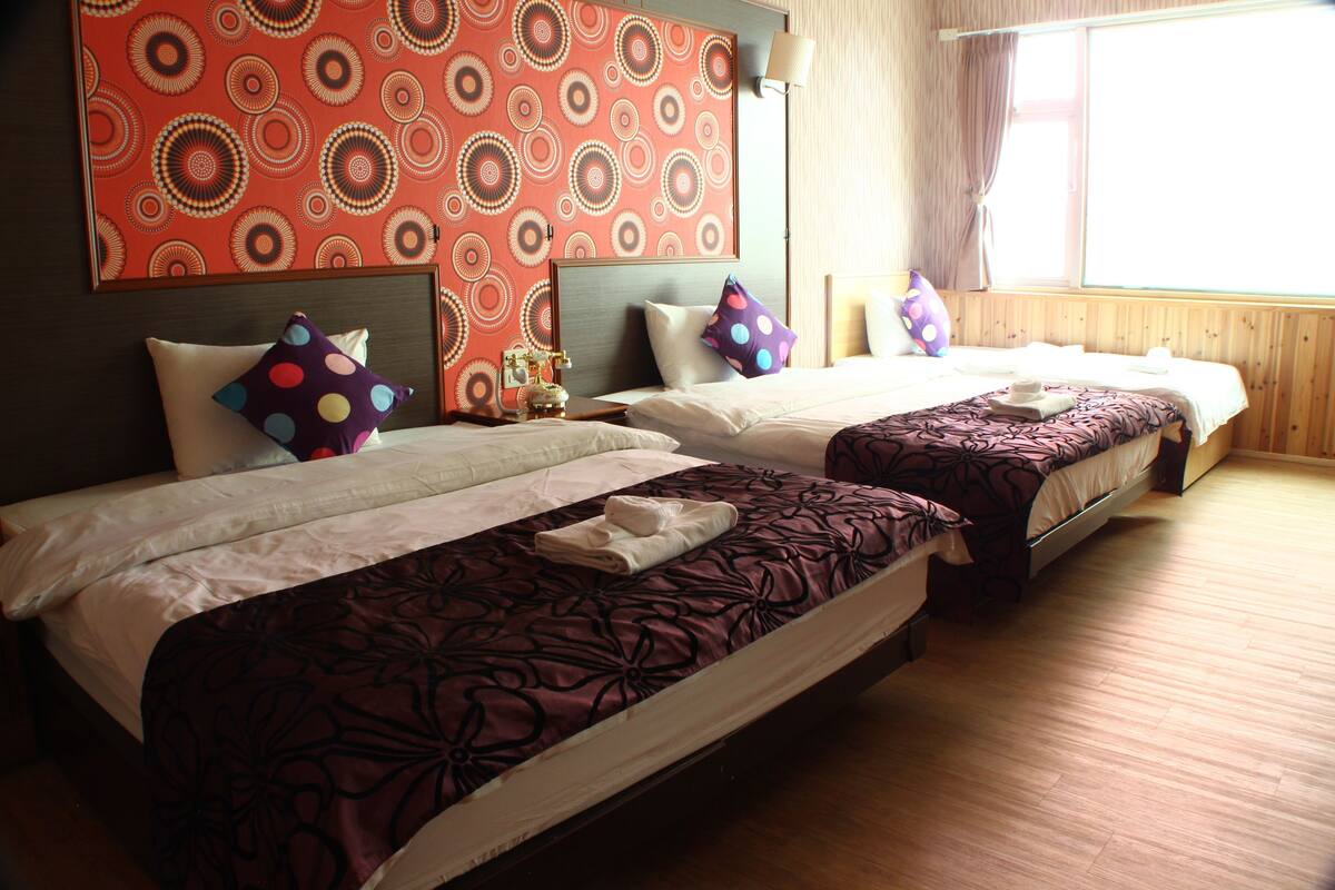 A hotel room with two beds, a patterned wall, wooden flooring, and a window with curtains.