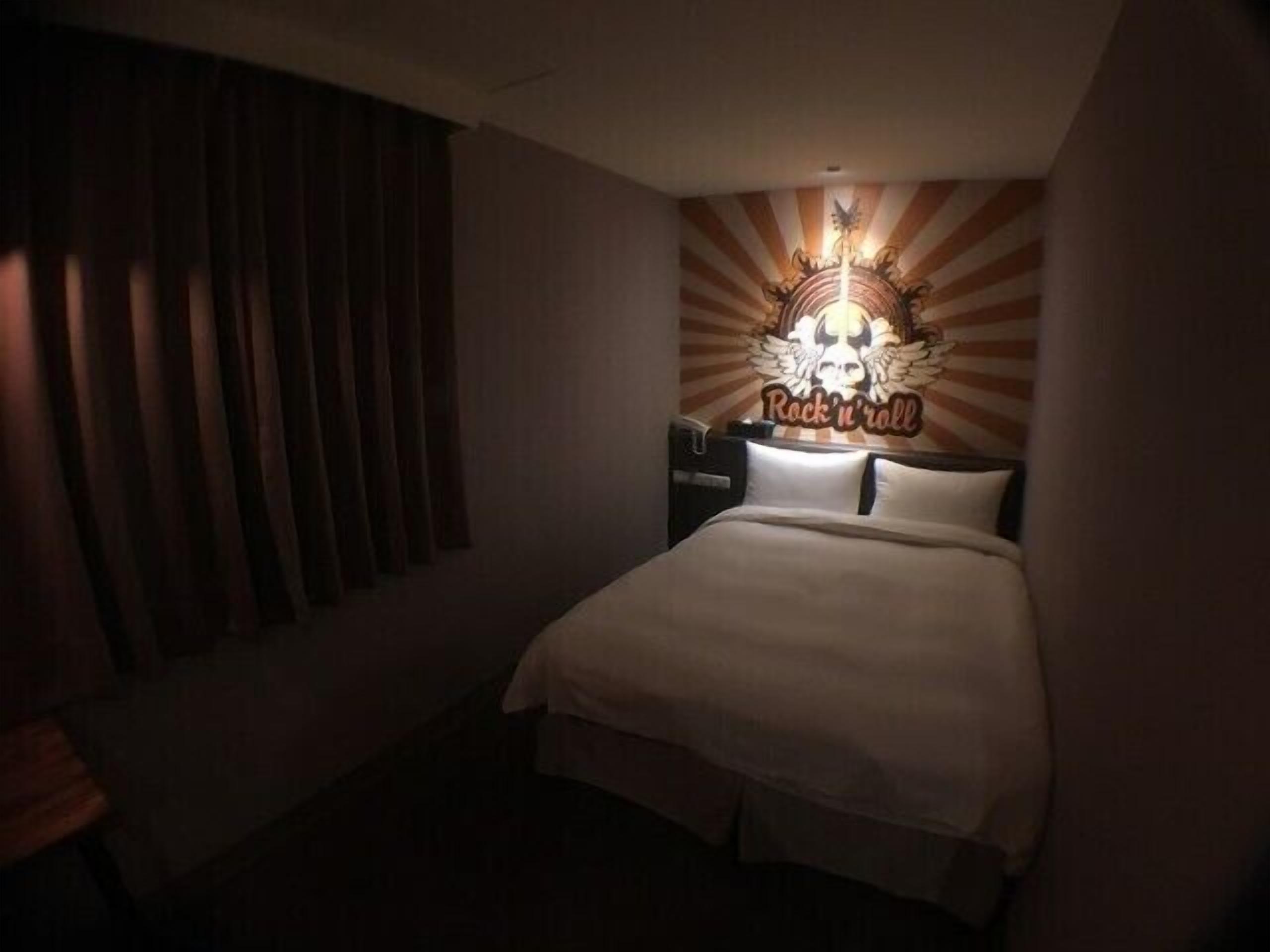 A bedroom with a bed, a nightstand, a wall-mounted lamp, and a window with blinds.