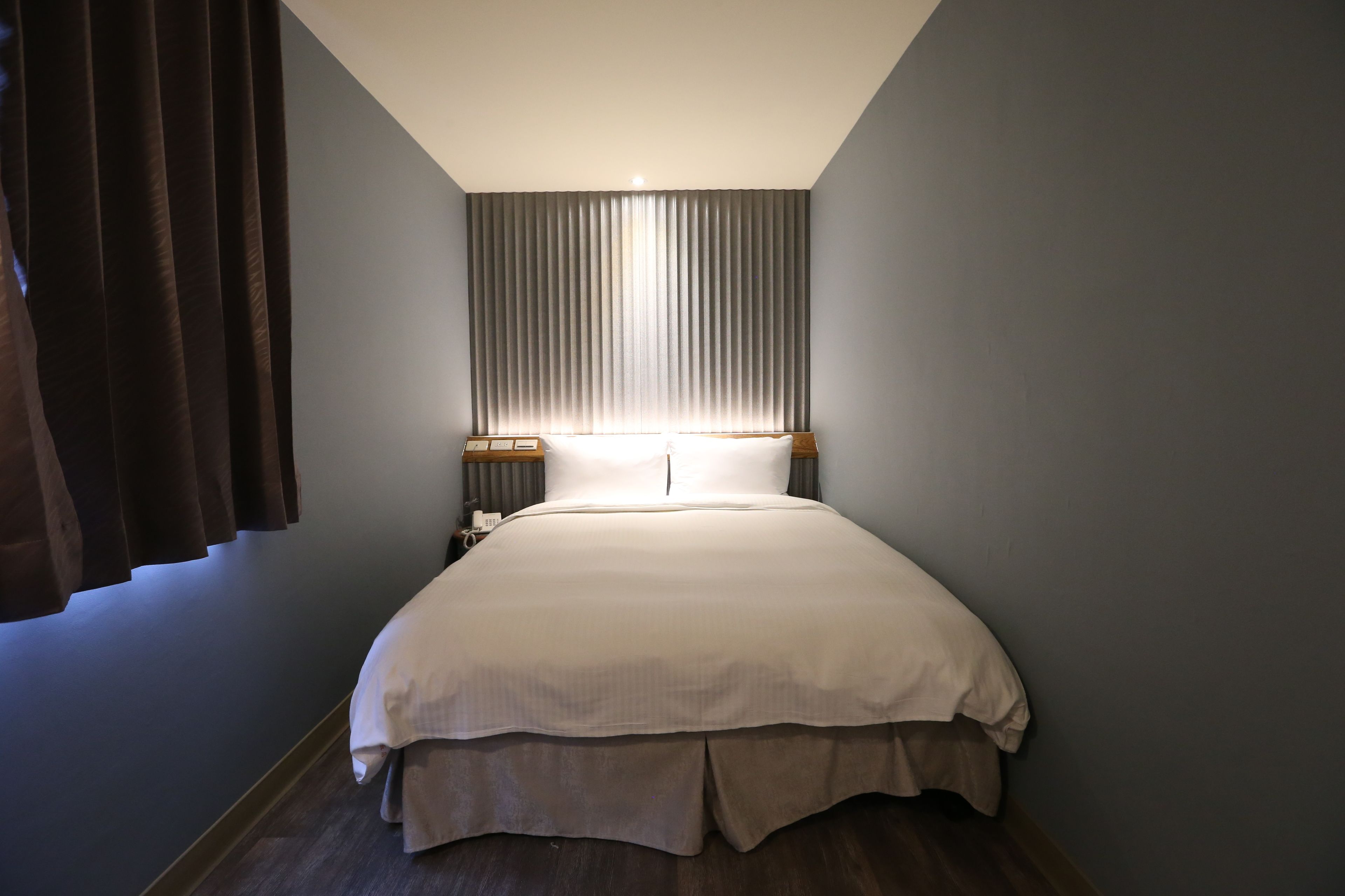 Una cama bien tendida en una habitación con un ventanal grande cubierto por persianas verticales, una mesita de noche con una lámpara y un aplique en la pared.