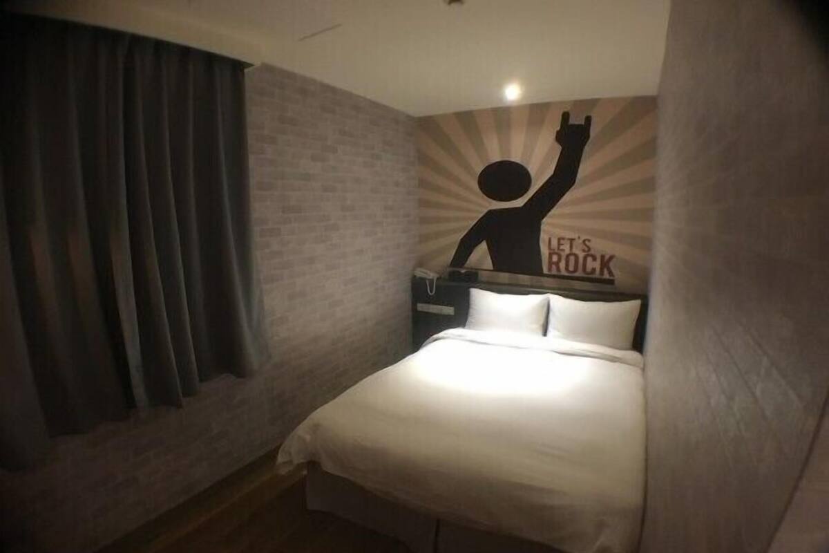 Una cama pequeña y bien hecha en una habitación de esquina con pared de ladrillo y un letrero que dice 'Let's Rock'.