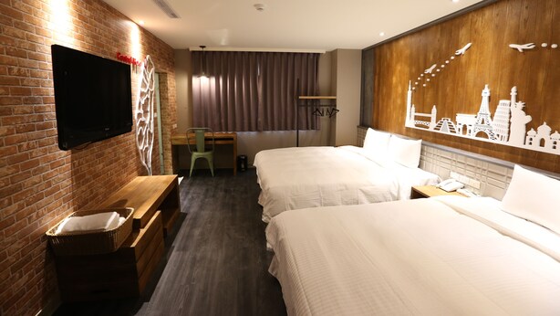 A hotel room with two beds, a flat-screen TV mounted on a brick wall, a wooden headboard with a cityscape mural, and a desk with a chair.