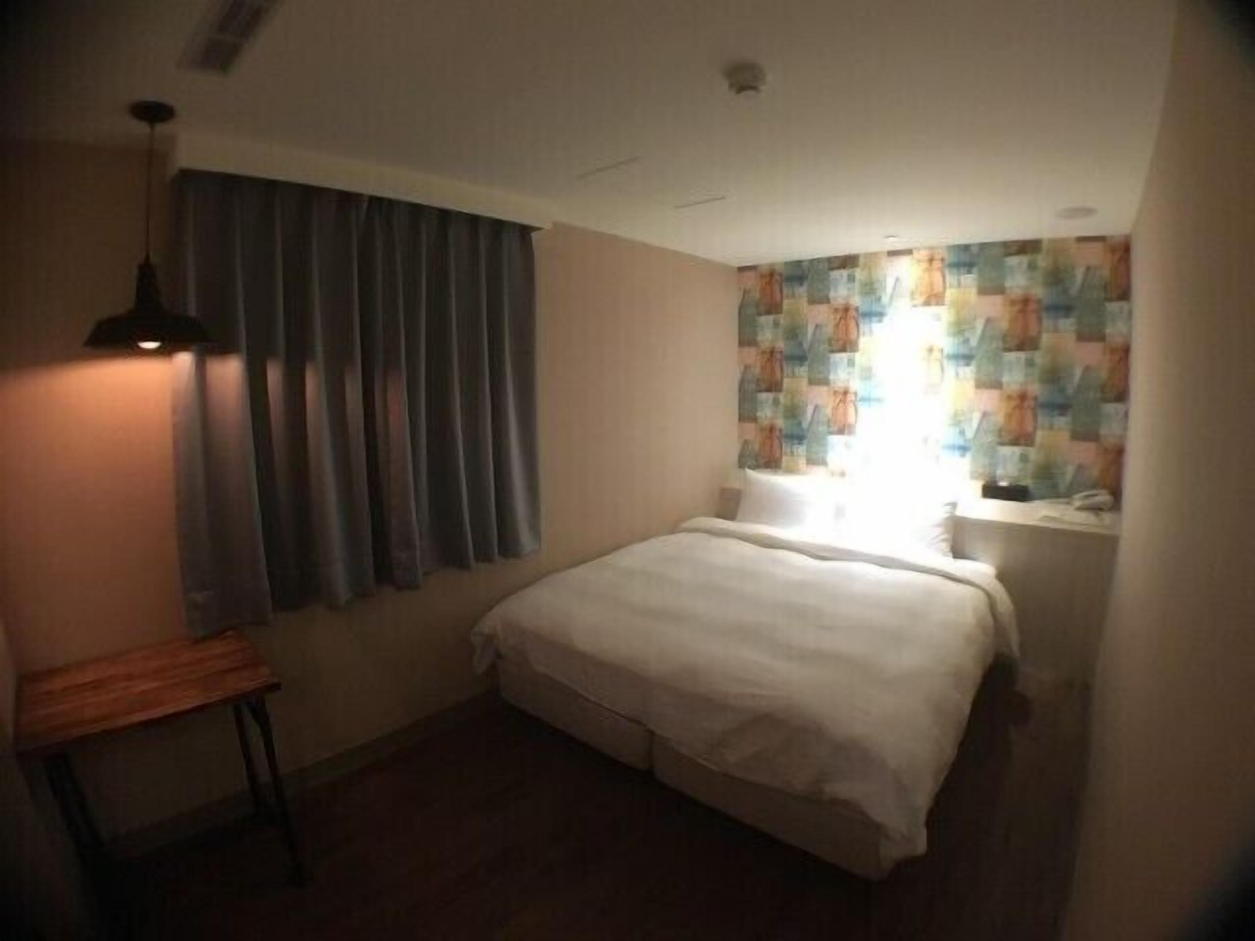 A small, neatly arranged bedroom with a bed, a bedside table, a lamp, and a window with curtains.