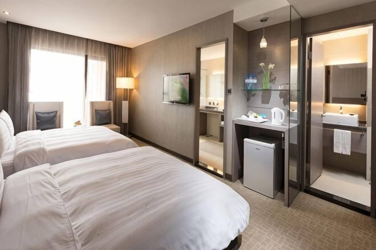 A hotel room with two beds, a desk with a mirror, a small kitchenette, and a bathroom with a sink and mirror.