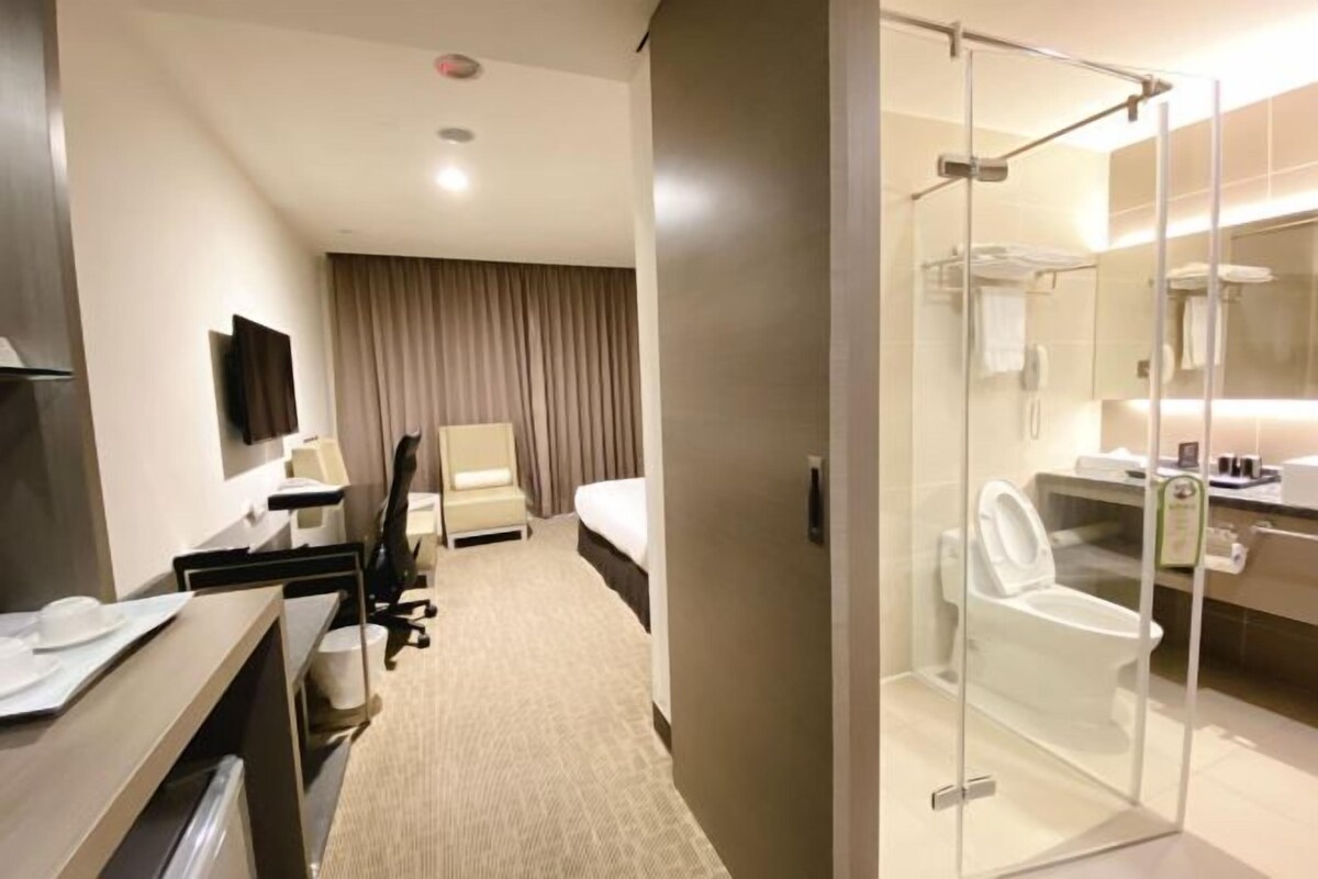 Quarto de hotel com cama, escrivaninha com cadeira, televisão, chuveiro com divisória de vidro e banheiro com vaso sanitário e pia.