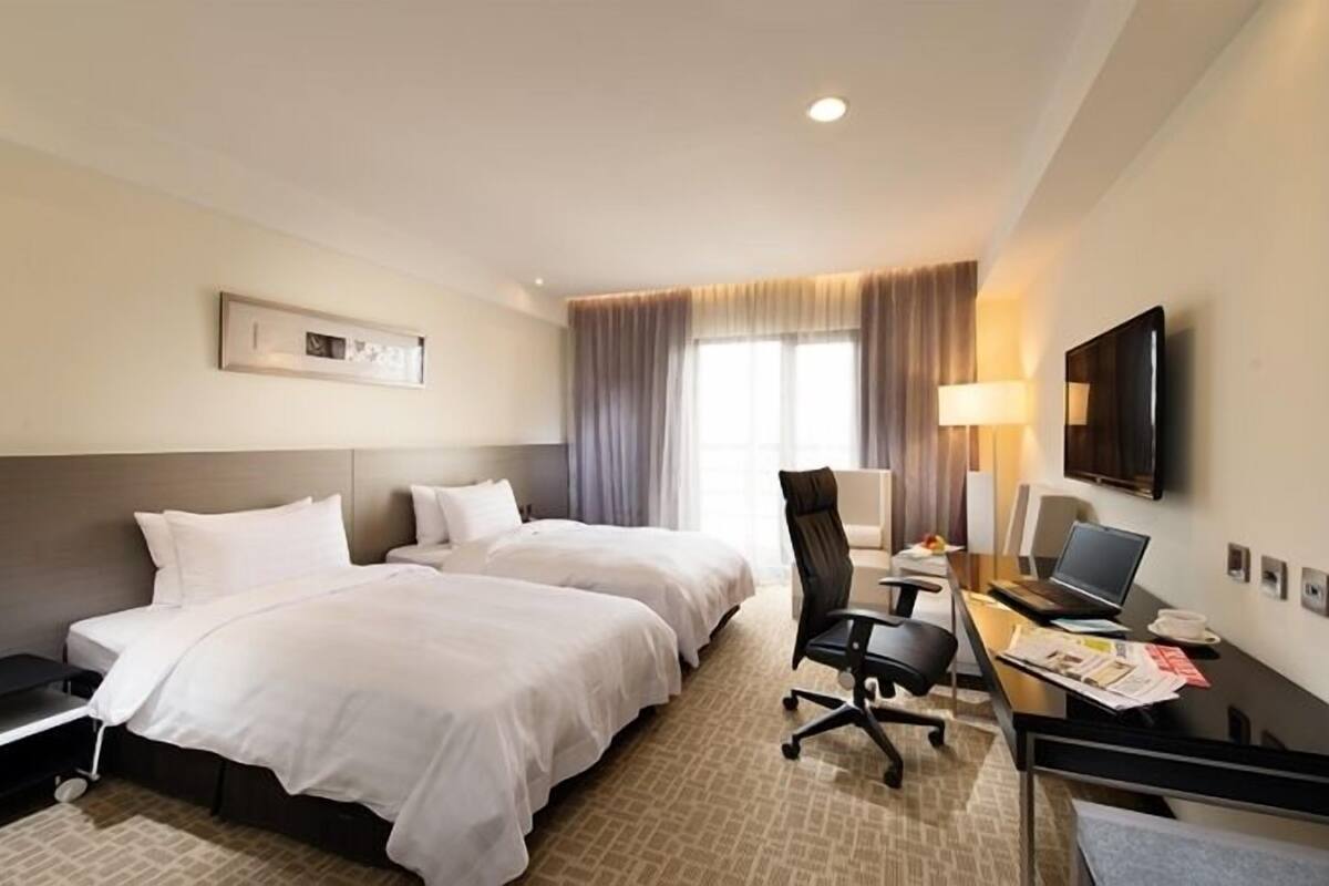 A hotel room with two beds, a desk with a chair, a flat-screen TV, and a window with curtains.