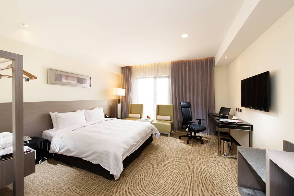 A hotel room with a large bed, a desk, a chair, a TV, and a window with curtains.