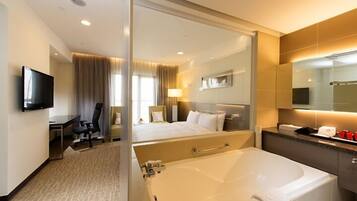 Habitación de hotel con una cama grande, zona de estar separada con un escritorio y una silla, televisor, bañera y espejo.