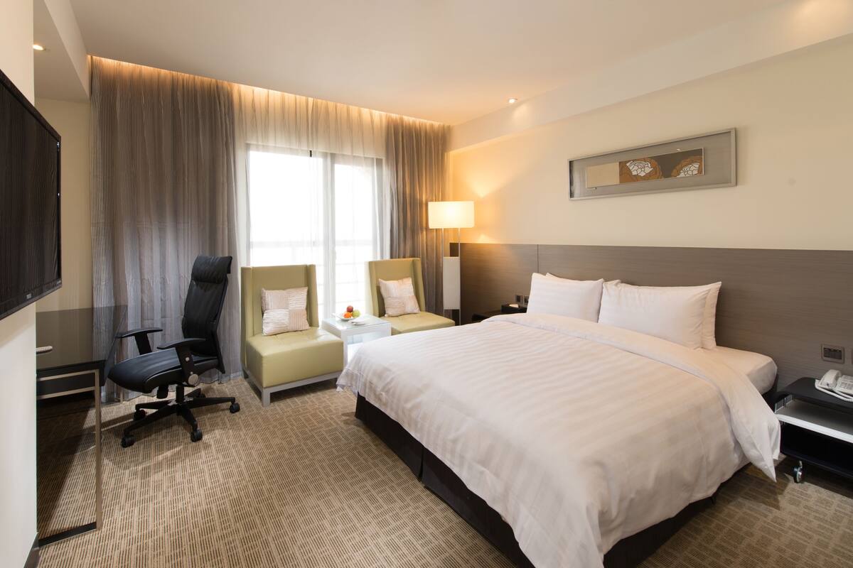 A hotel room with a large bed, two armchairs, a desk, a flat-screen TV, and a lamp.