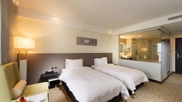 A hotel room with two beds, a desk with a fruit bowl, a lamp, and a glass-enclosed shower area.