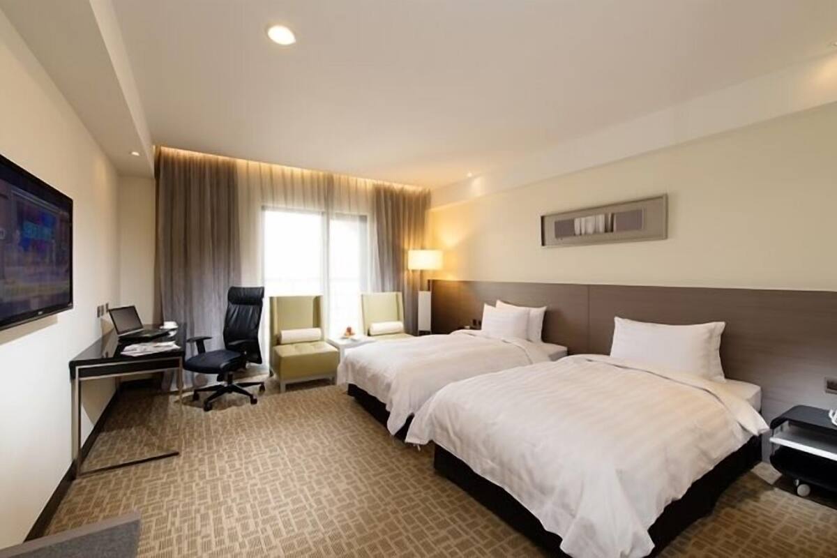 A hotel room with two beds, a desk with a computer, a chair, a TV, and a window with curtains.