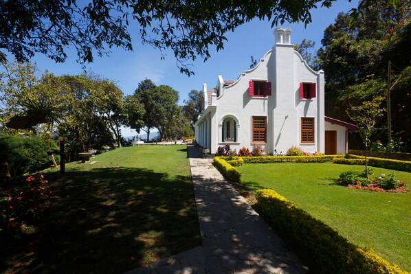 Un edificio blanco con contraventanas rojas, un sendero de piedra y jardines bien cuidados.