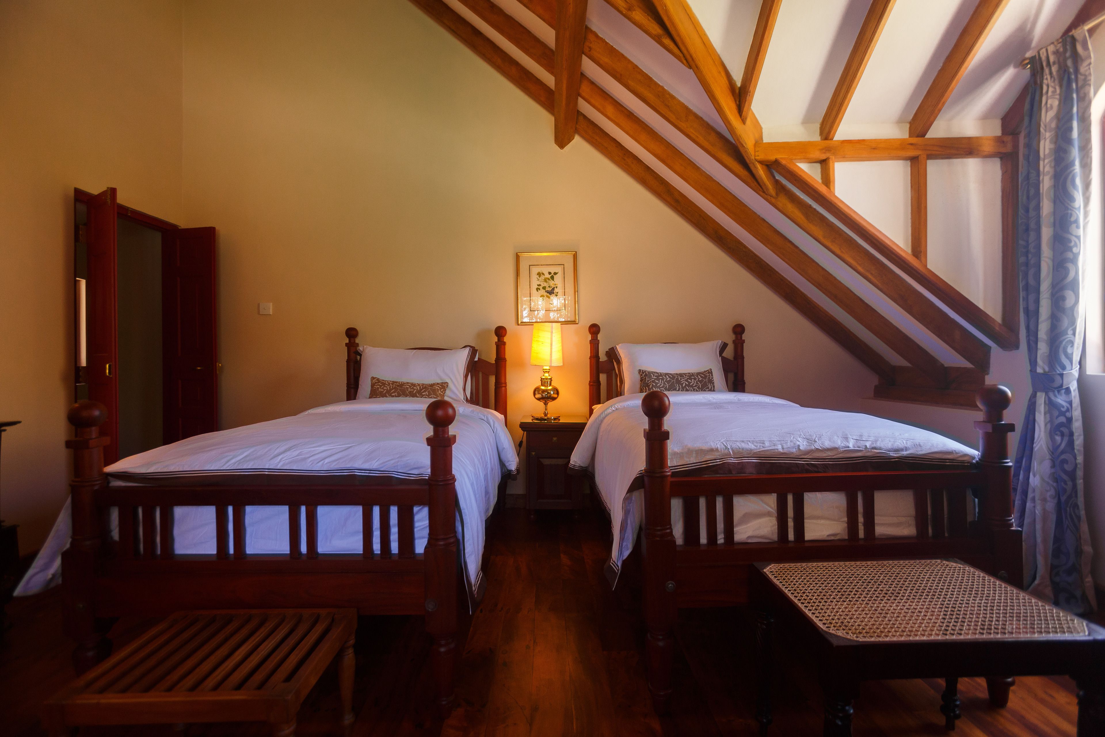 A bedroom with two wooden beds, a bench, a nightstand with a lamp, and a framed picture on the wall.