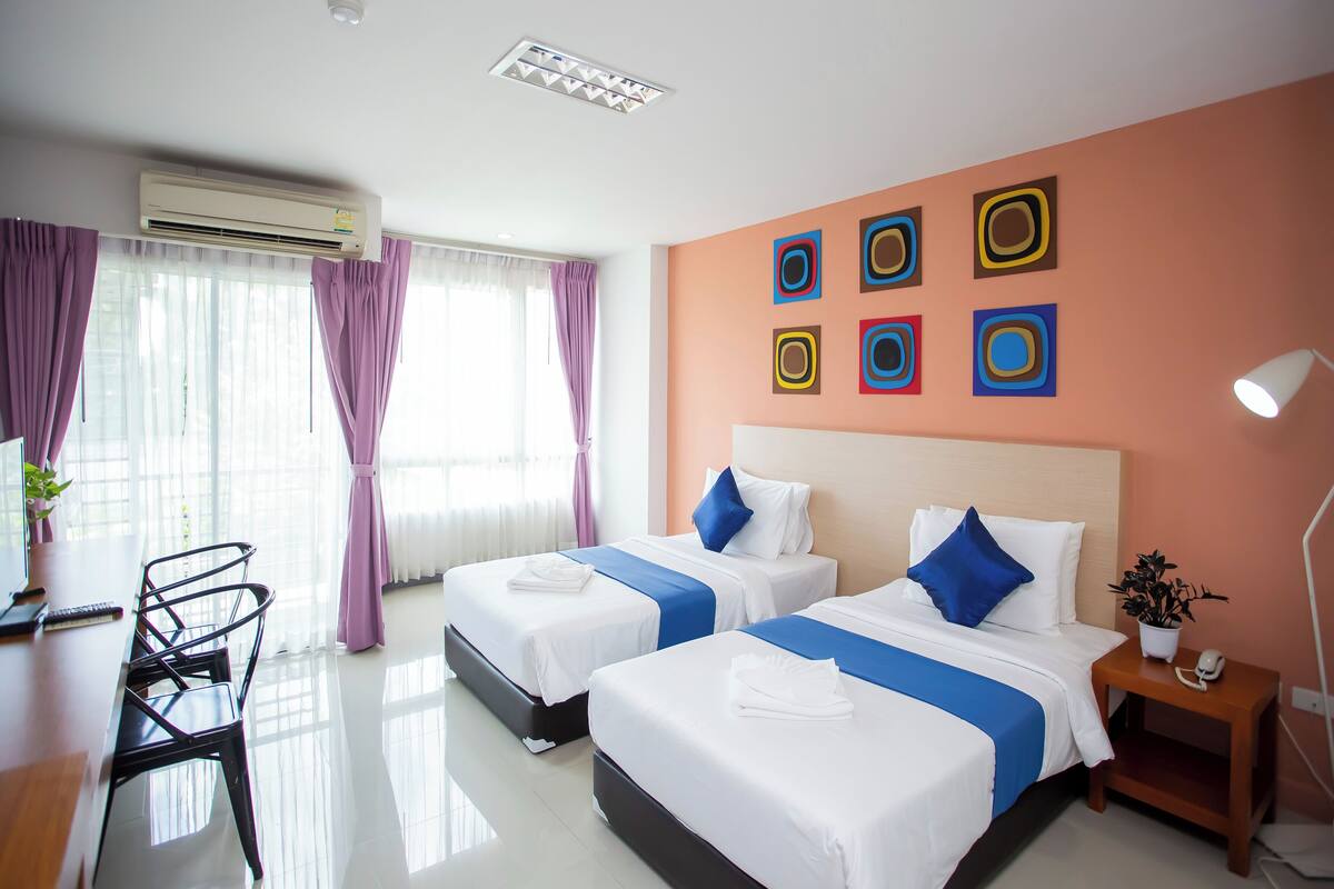 Une chambre d’hôtel avec deux lits, un bureau, une chaise, une table de chevet avec une lampe, et un mur orné d’œuvres d’art géométriques aux couleurs vives.