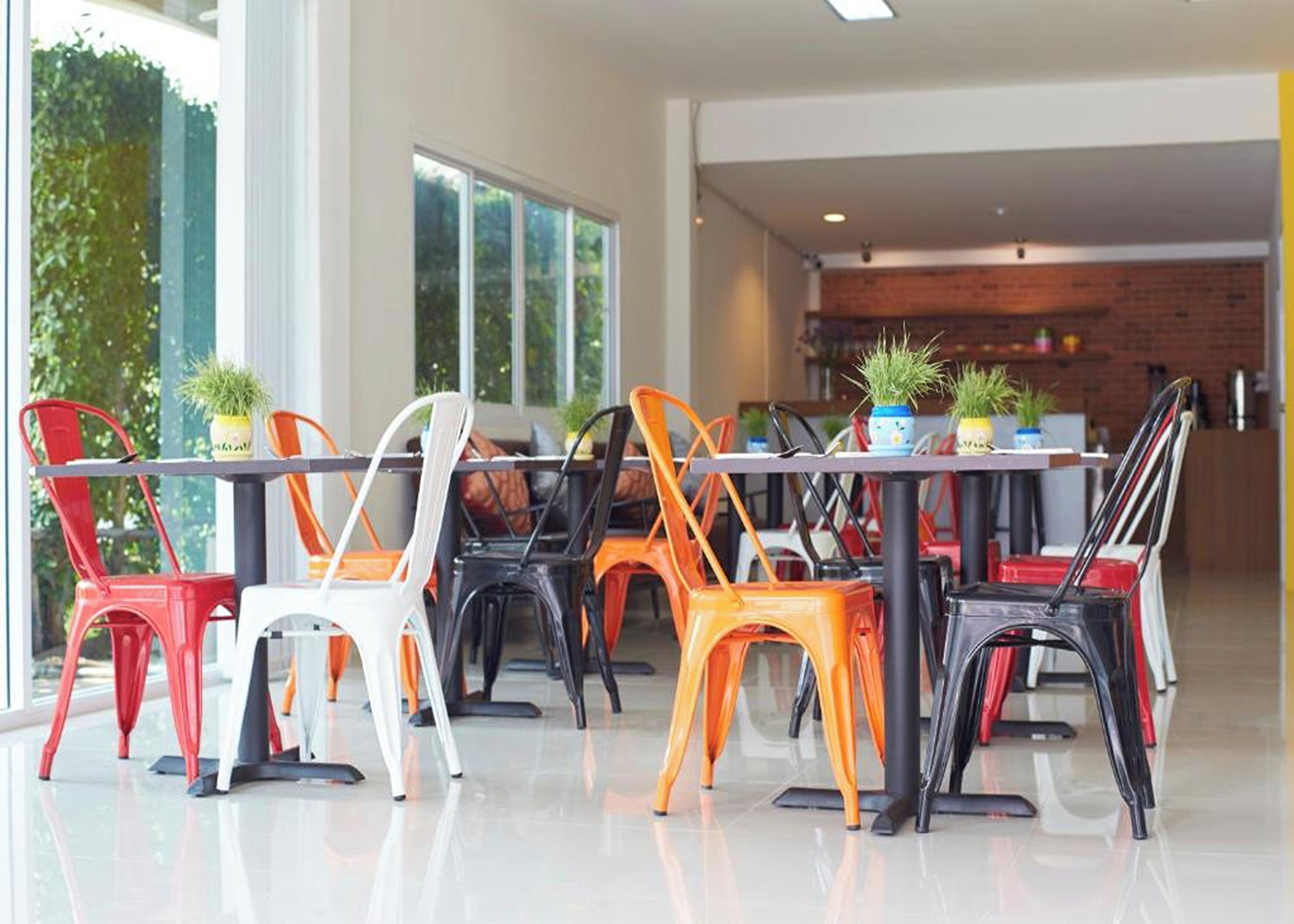 A modern dining area with a long table and colorful chairs, large windows, and a brick wall in the background.