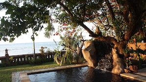 A swimming pool with a waterfall feature, surrounded by lush greenery and a view of the sea. - Joglo on the Beach (Banjar)