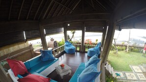 A covered outdoor seating area with blue and red cushions, a small table with flowers, and a view of the sea. - Joglo on the Beach (Banjar)