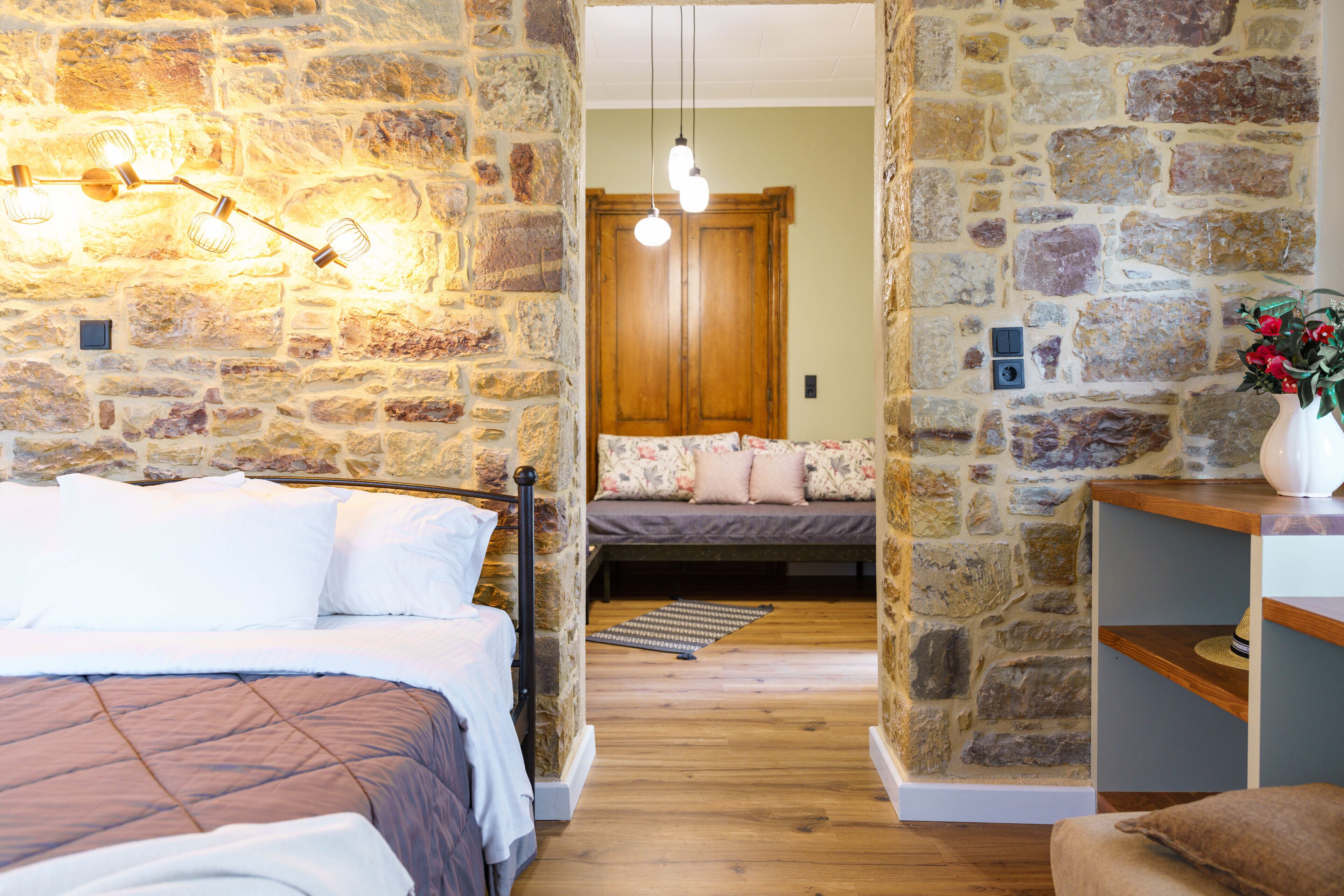 A cozy room with a stone wall, a bed with white and brown bedding, a wooden nightstand, and a vase with flowers.