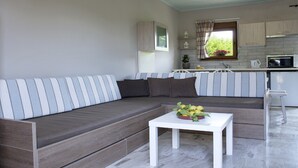 Un salon moderne avec un canapé à rayures grises, une table basse blanche agrémentée d’un arrangement floral et un coin cuisine doté de meubles en bois clair et d’une fenêtre avec des rideaux.