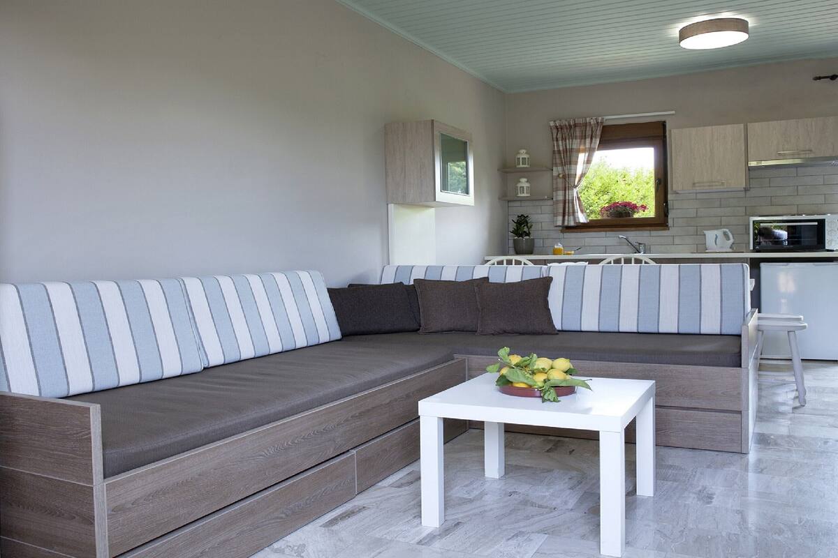 Una sala moderna con un sofá a rayas grises, una mesa de centro blanca con un arreglo floral, y una cocina con armarios de madera clara y una ventana con cortinas.