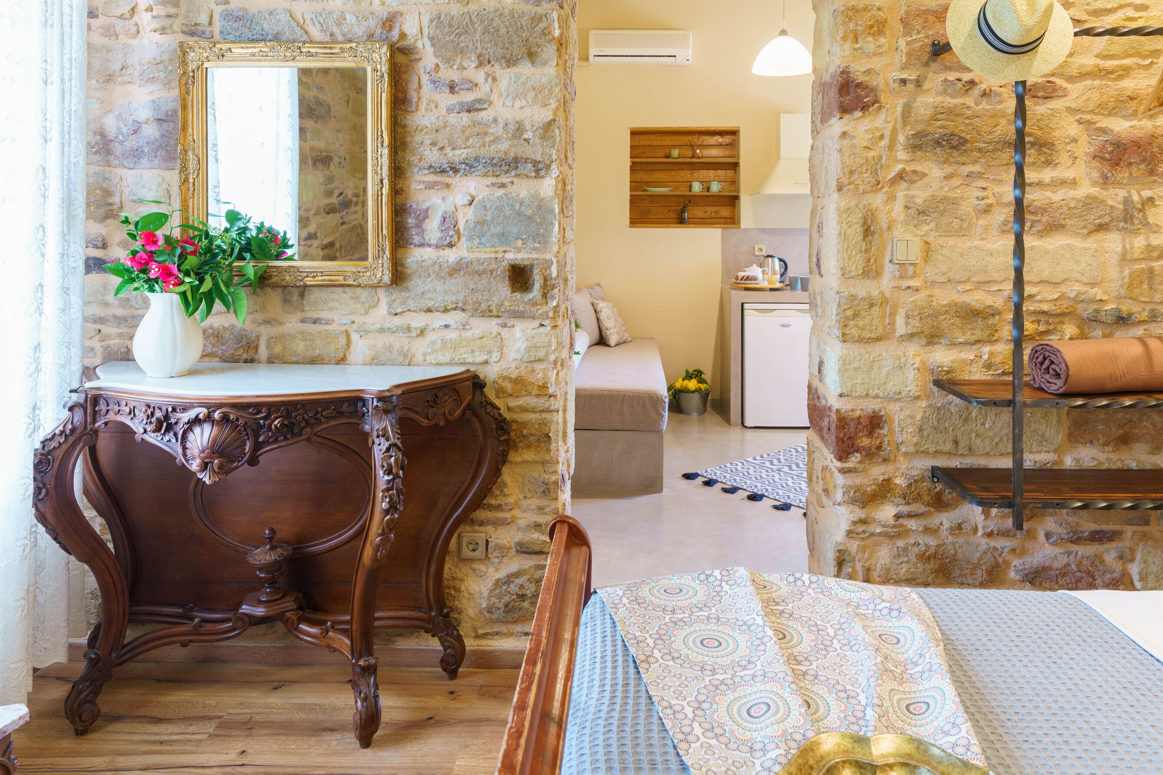 Una habitación con pared de piedra, una mesa de madera con un florero y una cama con ropa de cama estampada, además de un espejo en la pared.