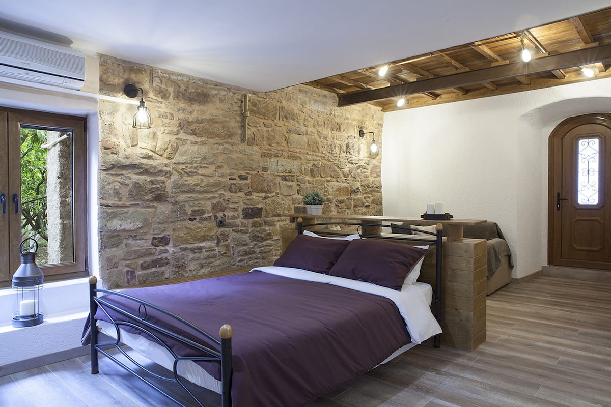 Un dormitorio con pared de piedra, techo de madera, cama con cabecera, ventana con vistas y una puerta.